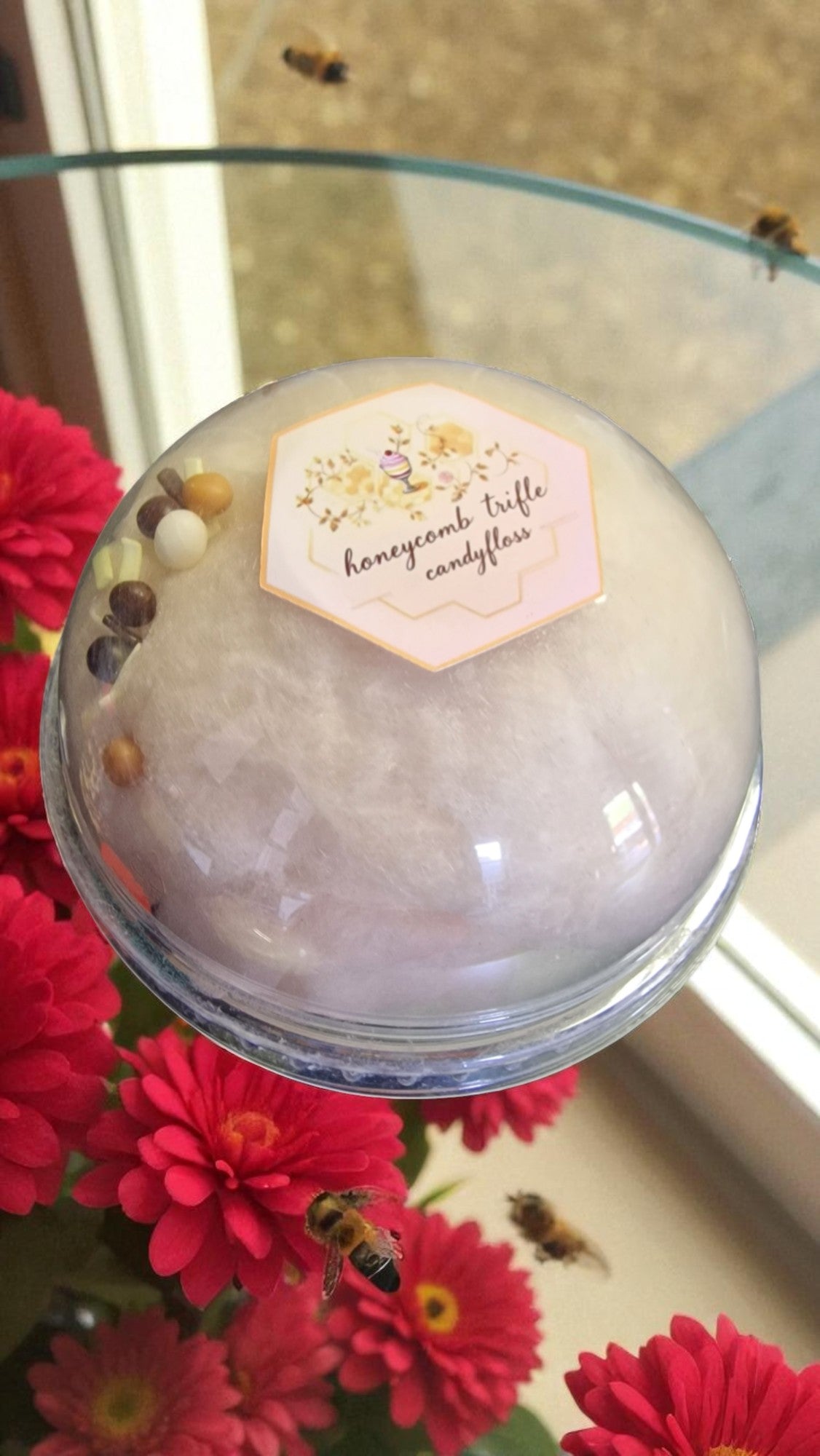 Honeycomb trifle candy ball with a label on a glass plate surrounded by red flowers and bees.