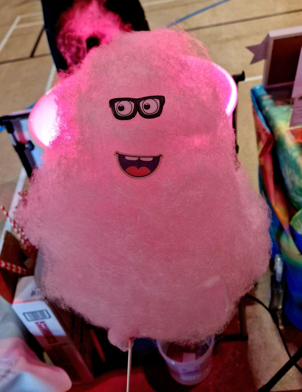 Pink cotton candy with a cartoon face and glasses on a stick, held by a person.
