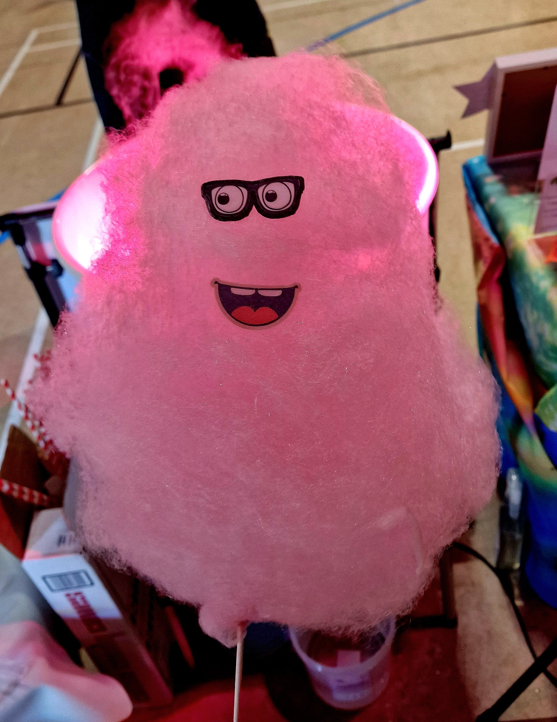 Pink cotton candy with a cartoon face and glasses on a stick, held by a person.