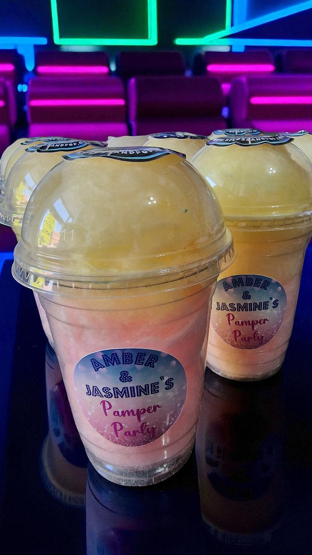 Two plastic cups with pink and yellow contents labeled 'Amber & Jasmine's Pamper Party' on a dark surface.