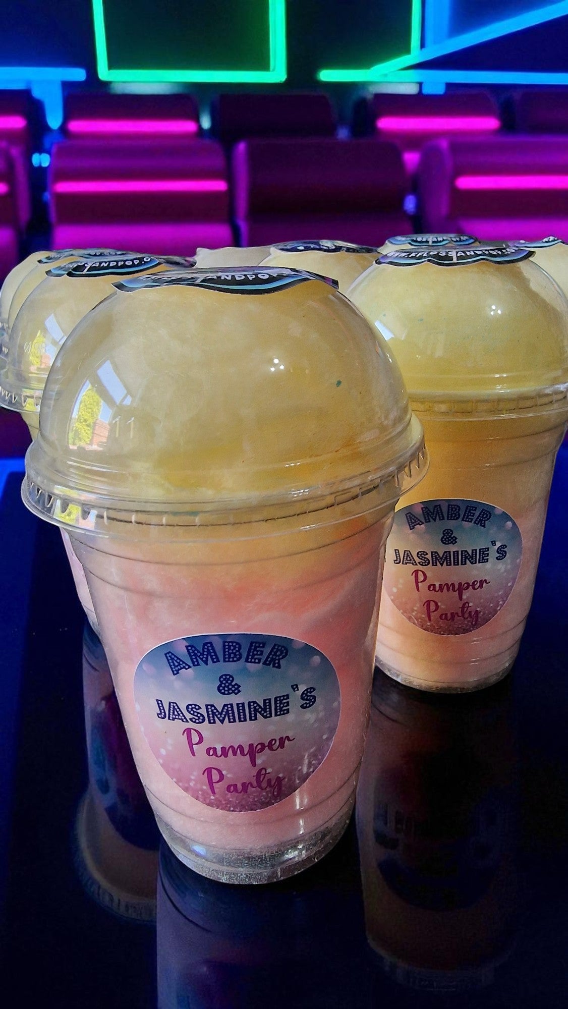Two plastic cups with pink and yellow contents labeled 'Amber & Jasmine's Pamper Party' on a dark surface.