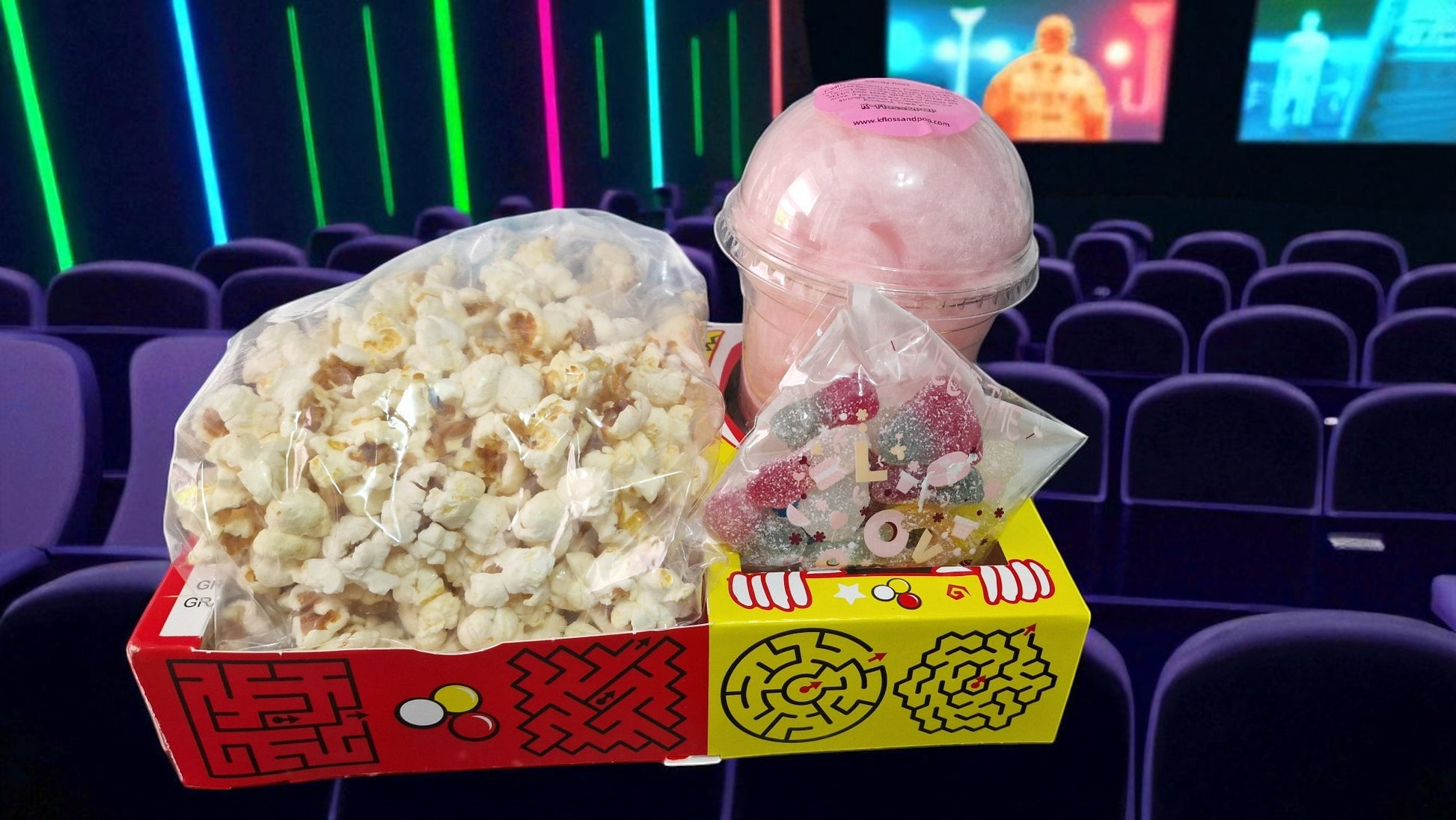 Popcorn, candy, and a drink in a theater setting with movie screens in the background.