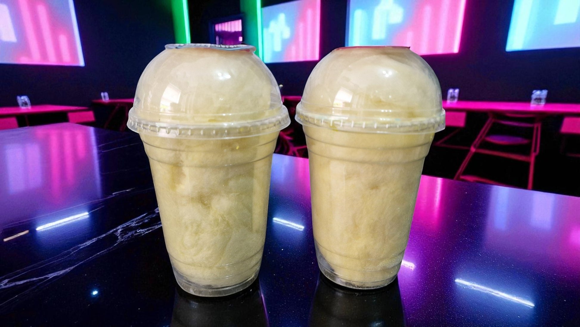 Two milkshakes in clear plastic cups with a colorful, blurred background