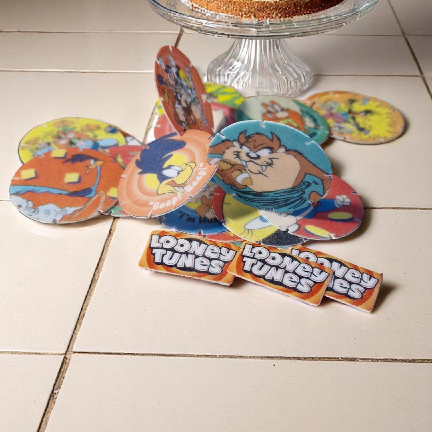 Colorful Looney Tunes coasters on a tiled floor with a glass cake stand in the background.