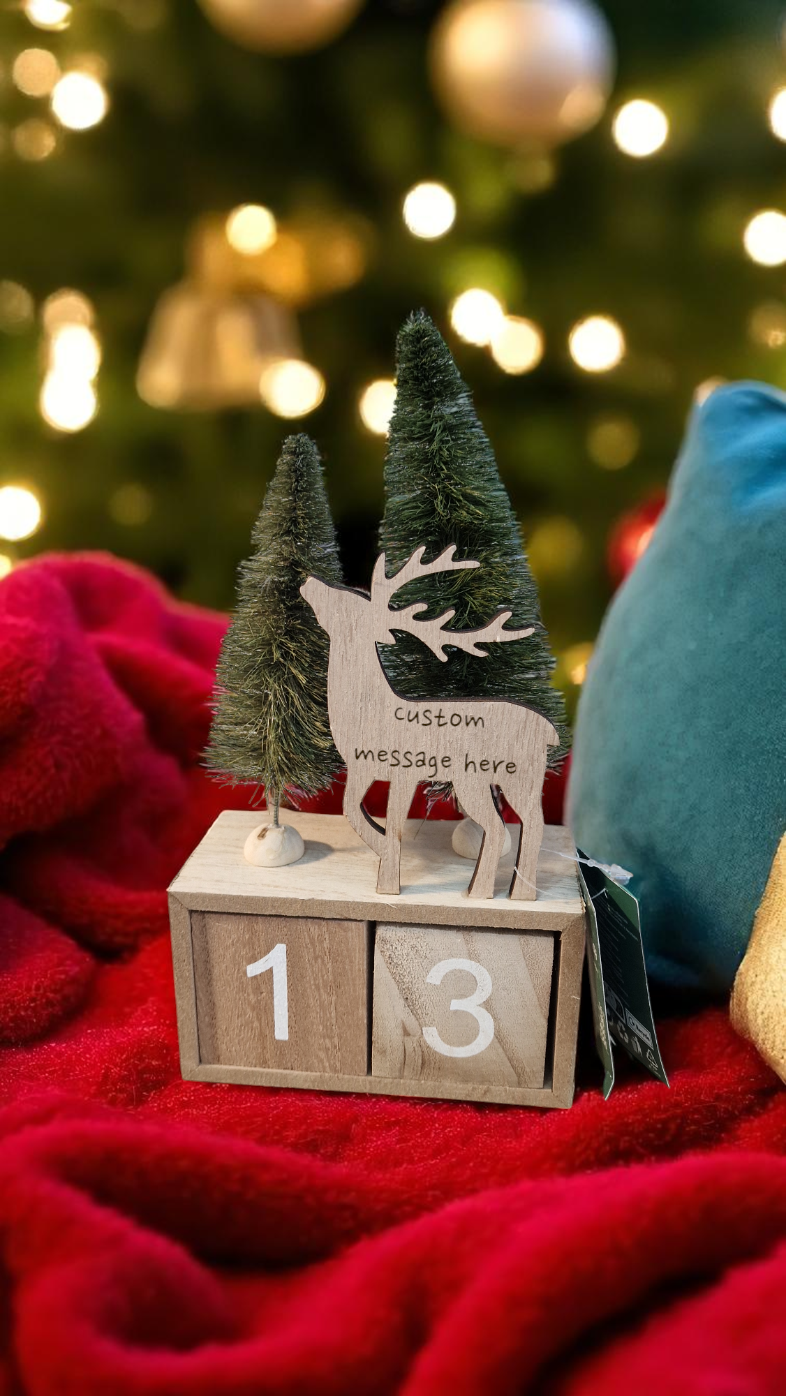 Decorative wooden calendar with a reindeer and trees on a red blanket with a blurred Christmas tree in the background.