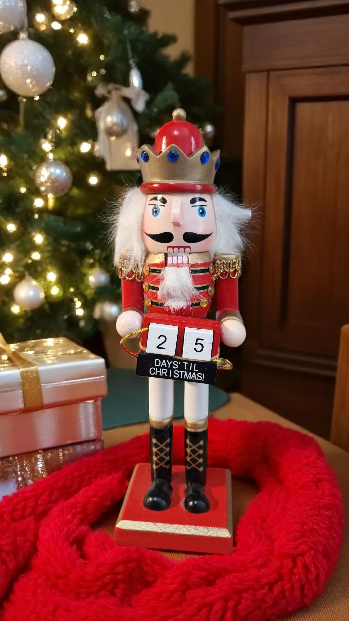 Red and gold wooden nutcracker advent calendar with white beard and hair, holding a gold staff, standing in front of a Christmas tree and gifts, displaying “25 days ‘til Christmas” on countdown blocks.