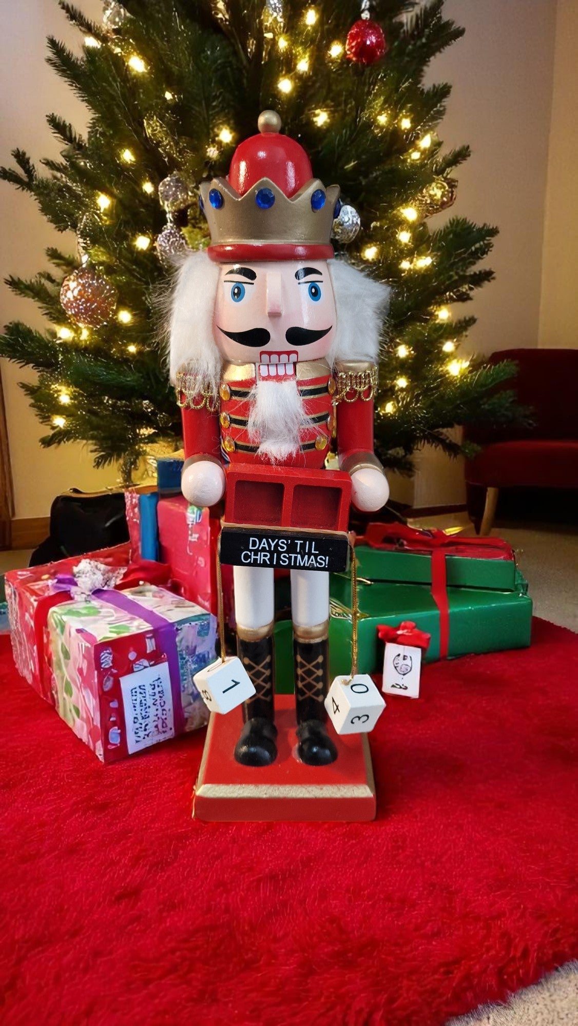 Red and gold wooden nutcracker advent calendar with white beard and hair, holding a gold staff, standing in front of a Christmas tree and gifts, displaying “25 days ‘til Christmas” on countdown blocks.
