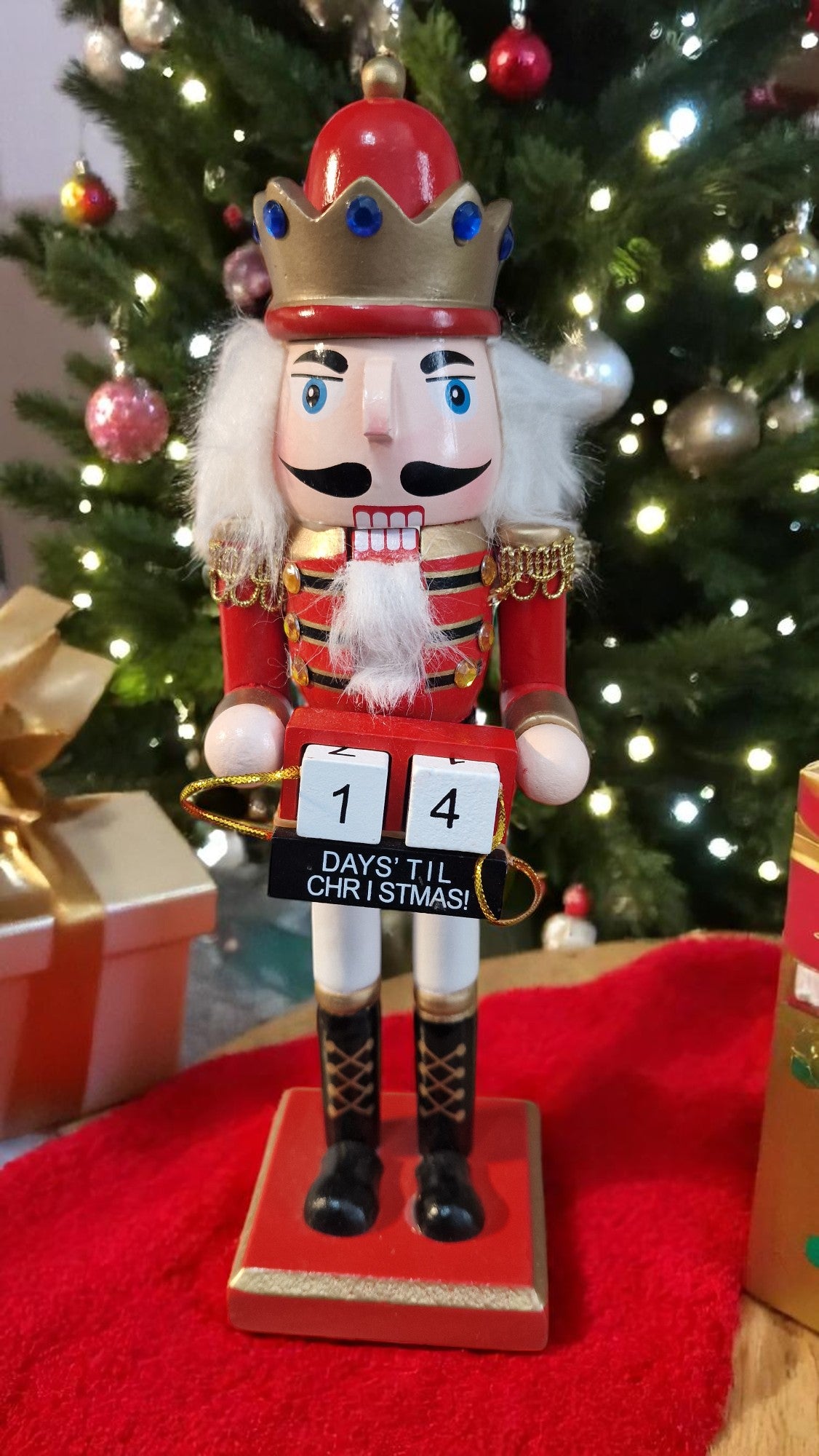 Red and gold wooden nutcracker advent calendar with white beard and hair, holding a gold staff, standing in front of a Christmas tree and gifts, displaying “25 days ‘til Christmas” on countdown blocks.