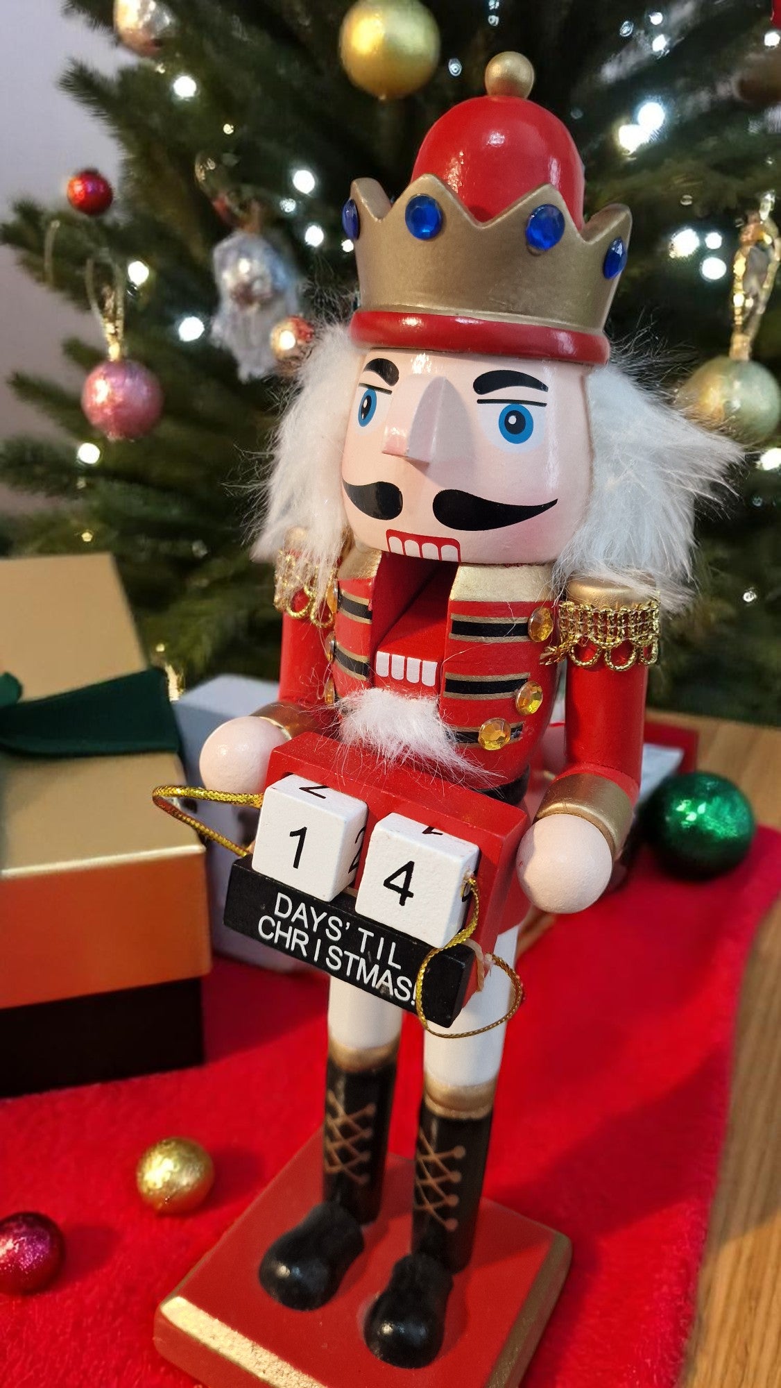 Red and gold wooden nutcracker advent calendar with white beard and hair, holding a gold staff, standing in front of a Christmas tree and gifts, displaying “25 days ‘til Christmas” on countdown blocks.