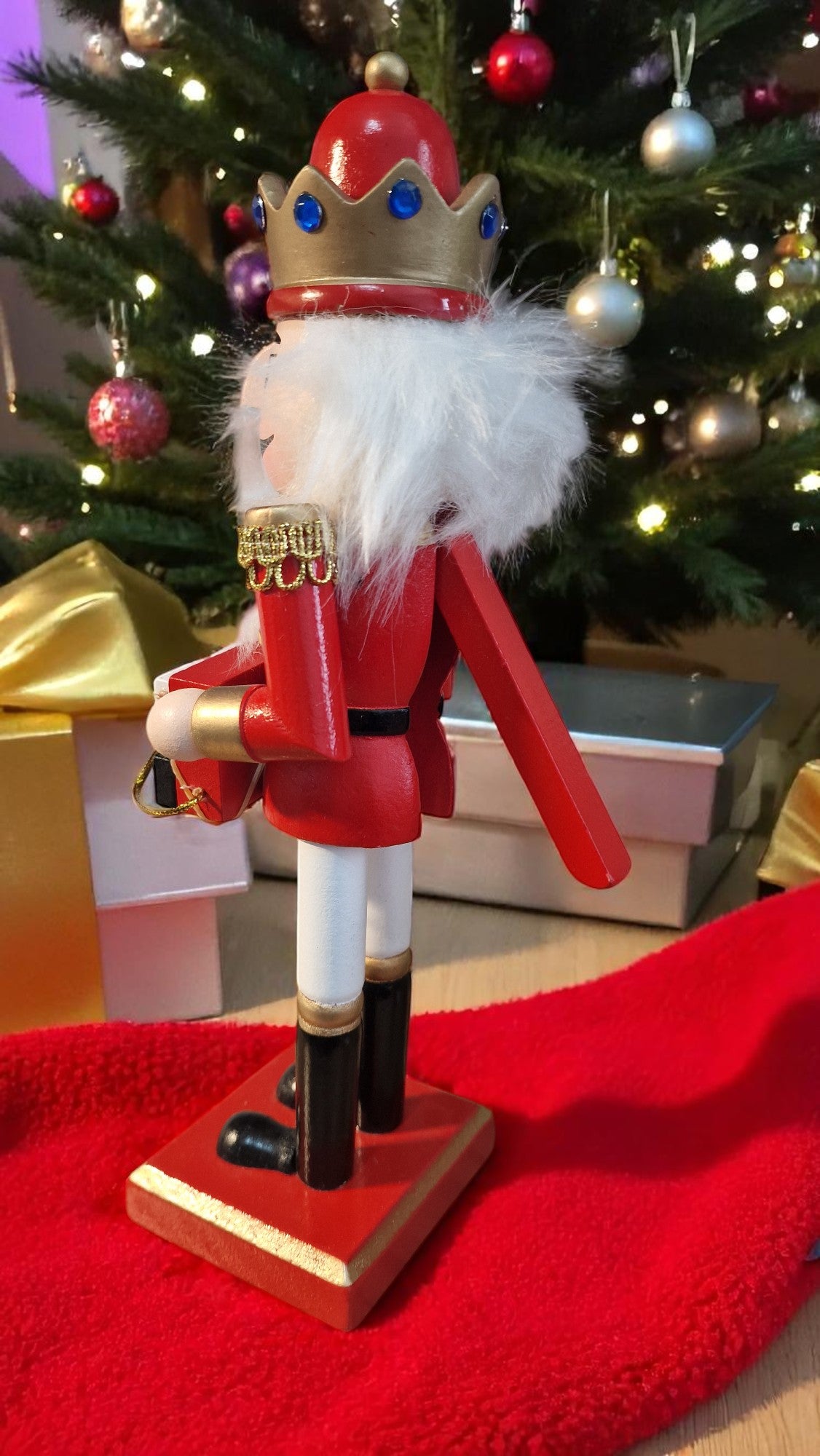 Red and gold wooden nutcracker advent calendar with white beard and hair, holding a gold staff, standing in front of a Christmas tree and gifts, displaying “25 days ‘til Christmas” on countdown blocks.