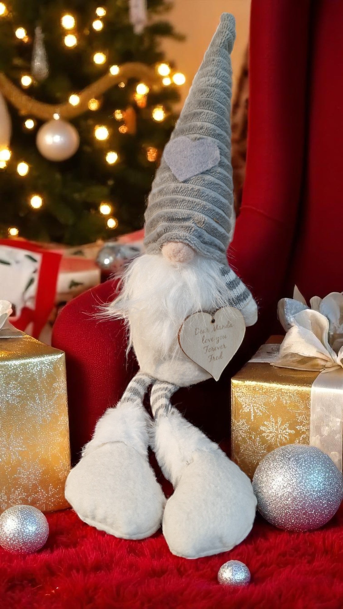 Decorative gnome with a heart tag in front of a Christmas tree and presents.