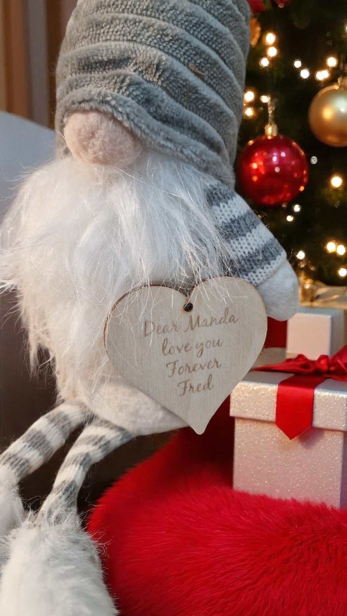 50cm Christmas gnome with red-and-white striped hat, white beard, and personalised wooden heart, sitting on a fireplace with festive lights in the background