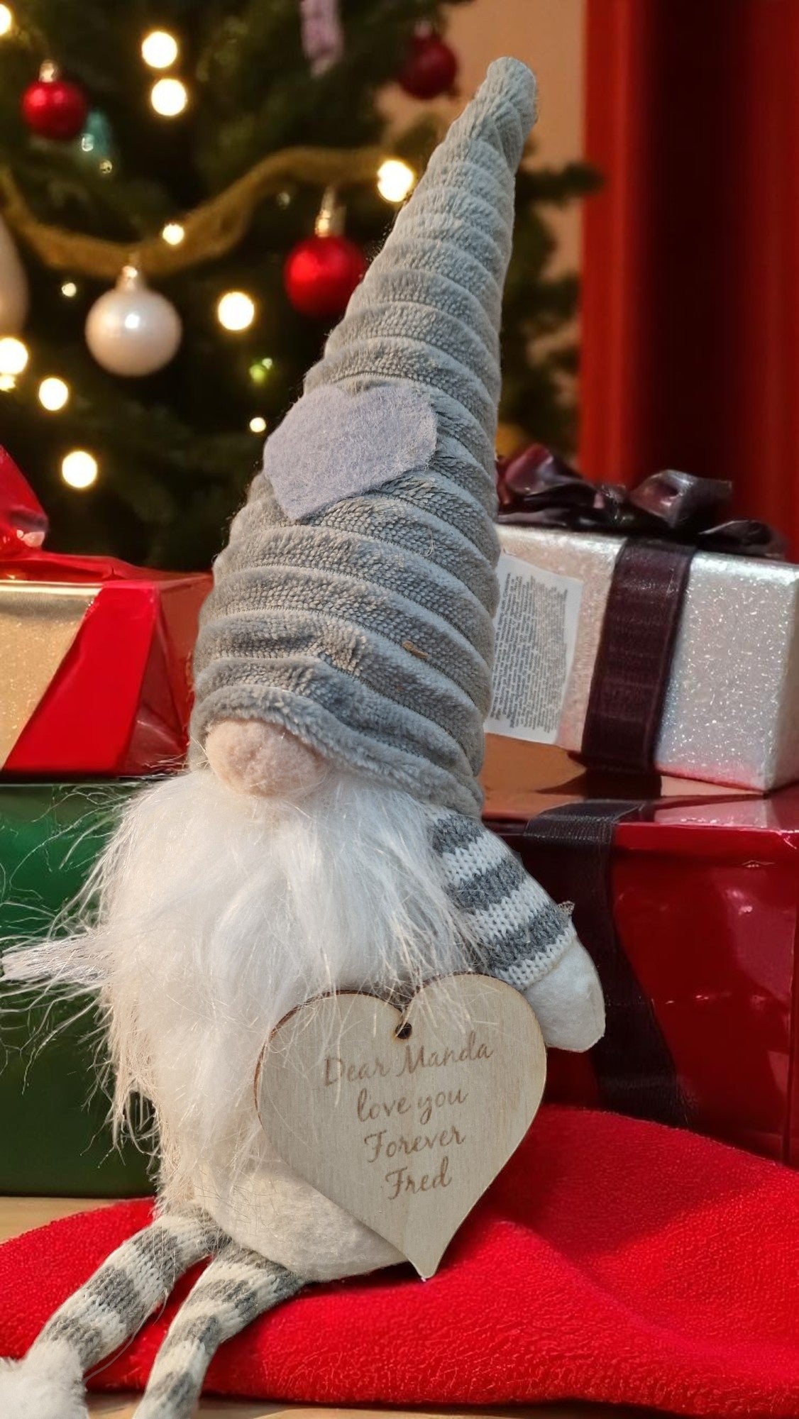 Christmas gnome with a heart-shaped sign in front of a decorated Christmas tree and presents.