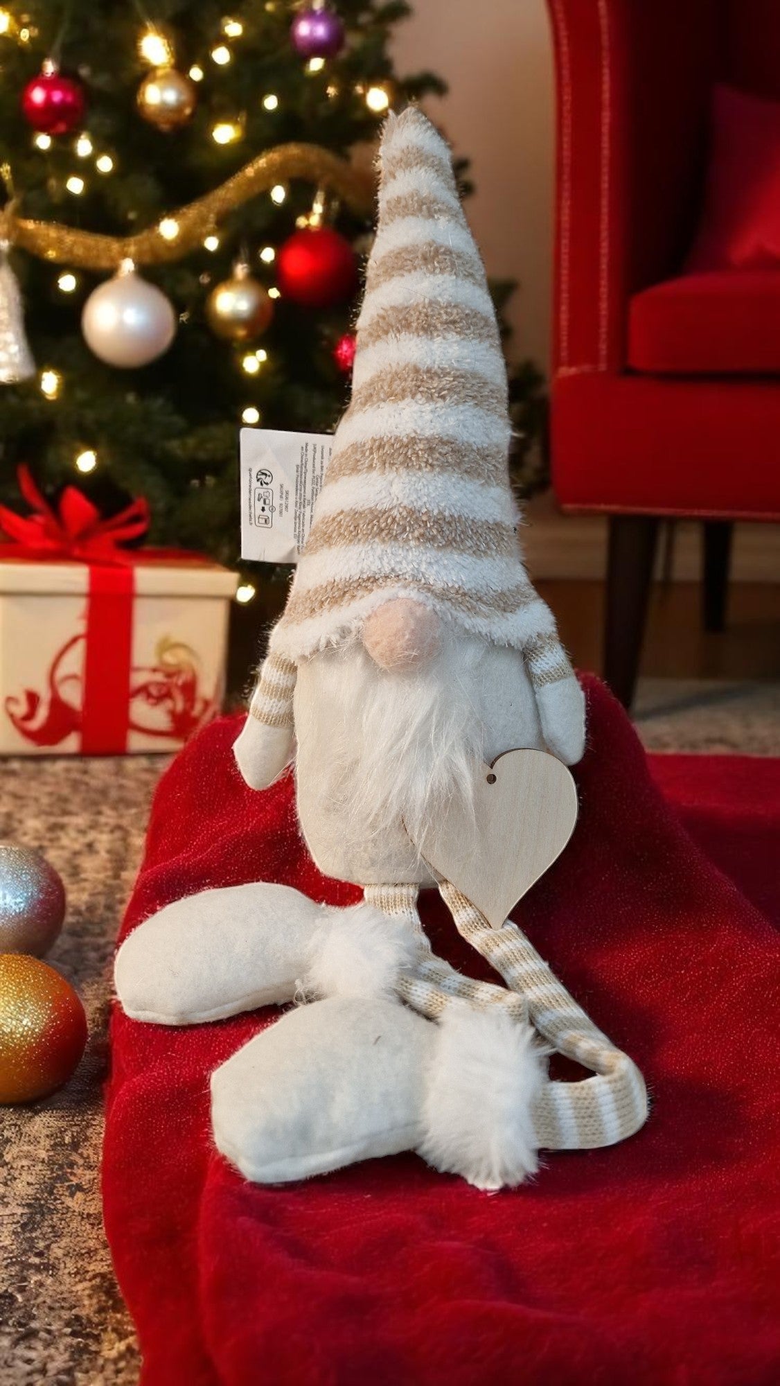 Decorative gnome with striped hat on a red surface, Christmas tree and gifts in the background