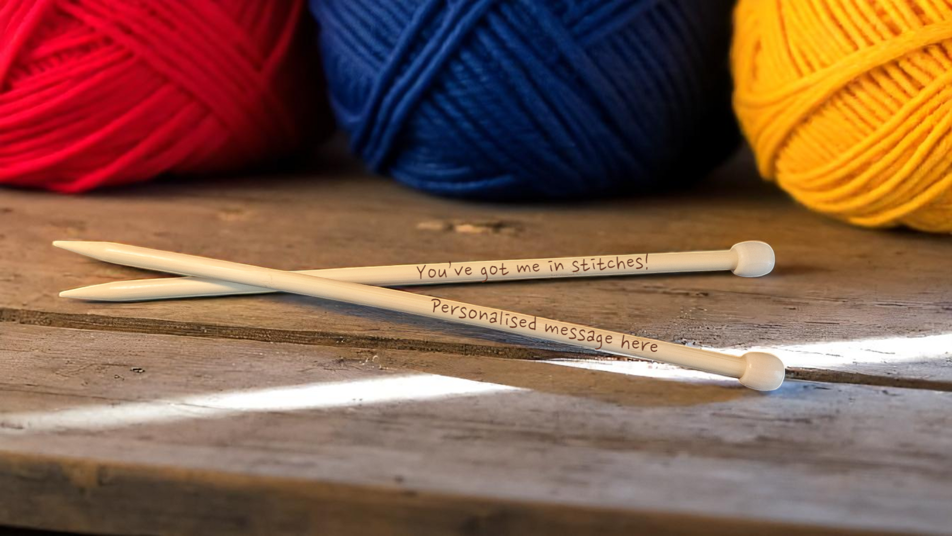 Pair of personalised bamboo knitting needles with engraved messages, chunky wool wrapped around, displayed on a wooden table with colourful yarn in the background