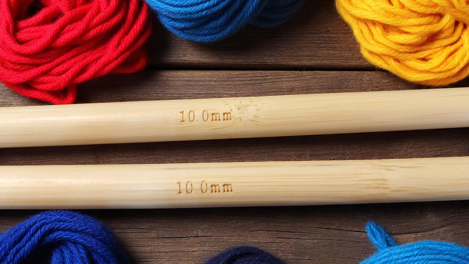 Pair of personalised bamboo knitting needles with engraved messages, chunky wool wrapped around, displayed on a wooden table with colourful yarn in the background