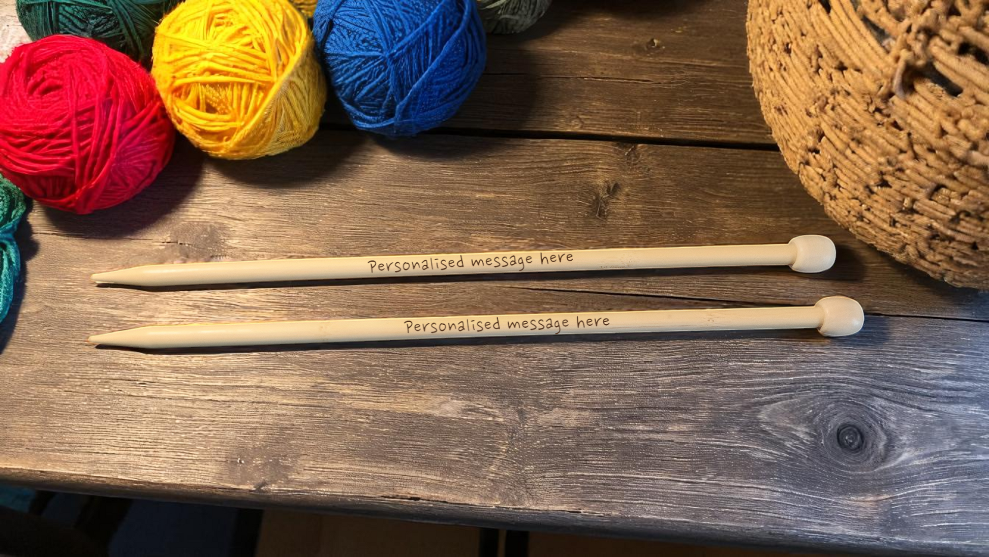 Pair of personalised bamboo knitting needles with engraved messages, chunky wool wrapped around, displayed on a wooden table with colourful yarn in the background