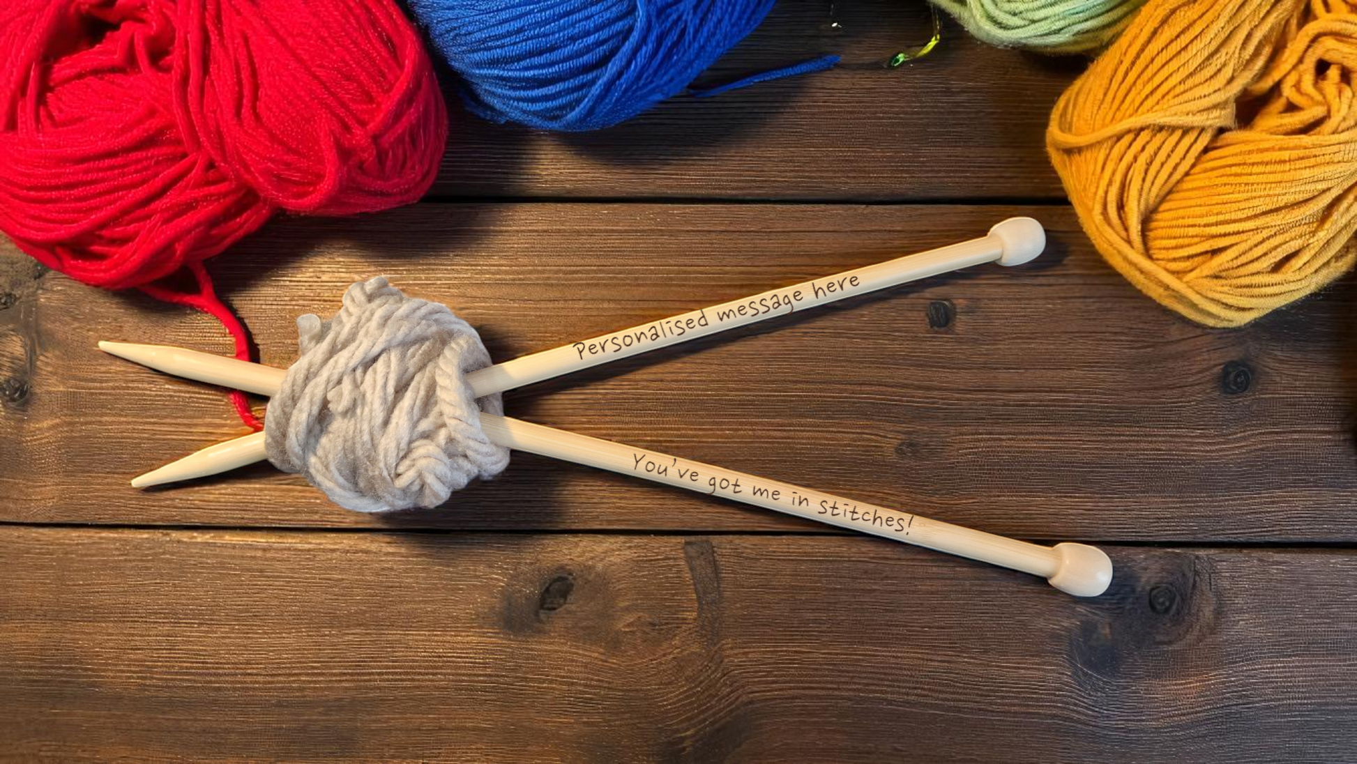 Pair of personalised bamboo knitting needles with engraved messages, chunky wool wrapped around, displayed on a wooden table with colourful yarn in the background