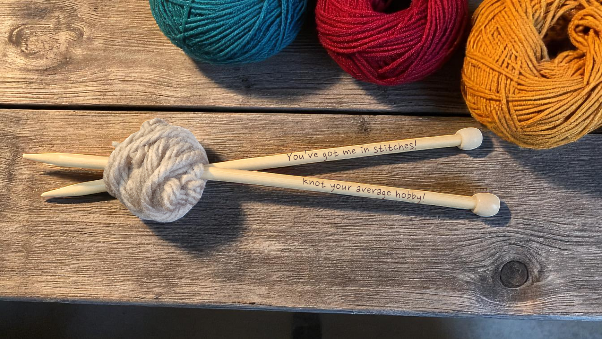 Pair of personalised bamboo knitting needles with engraved messages, chunky wool wrapped around, displayed on a wooden table with colourful yarn in the background