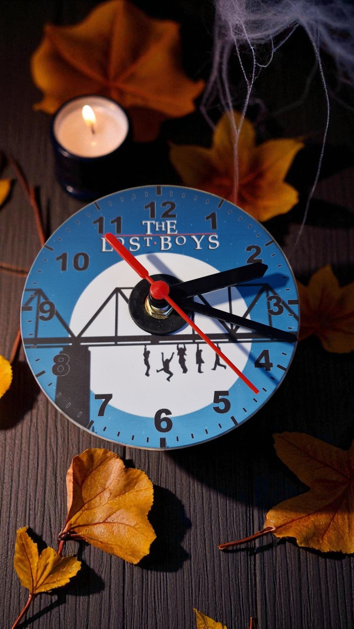 Wall clock with 'The Lost Boys' design on a dark surface with autumn leaves and a candle.
