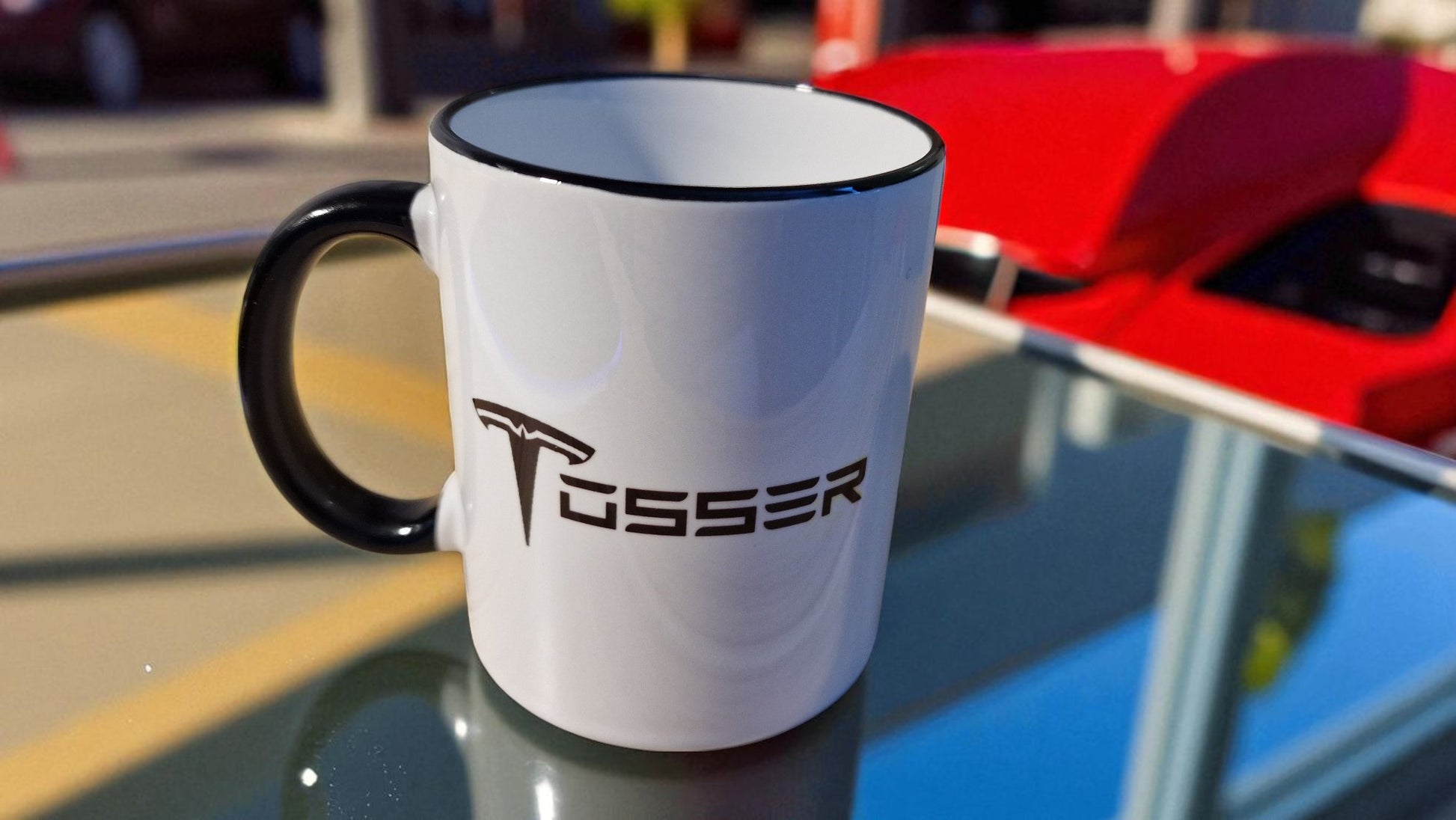 White mug with black interior and handle, featuring a logo, on a reflective surface with a blurred background.