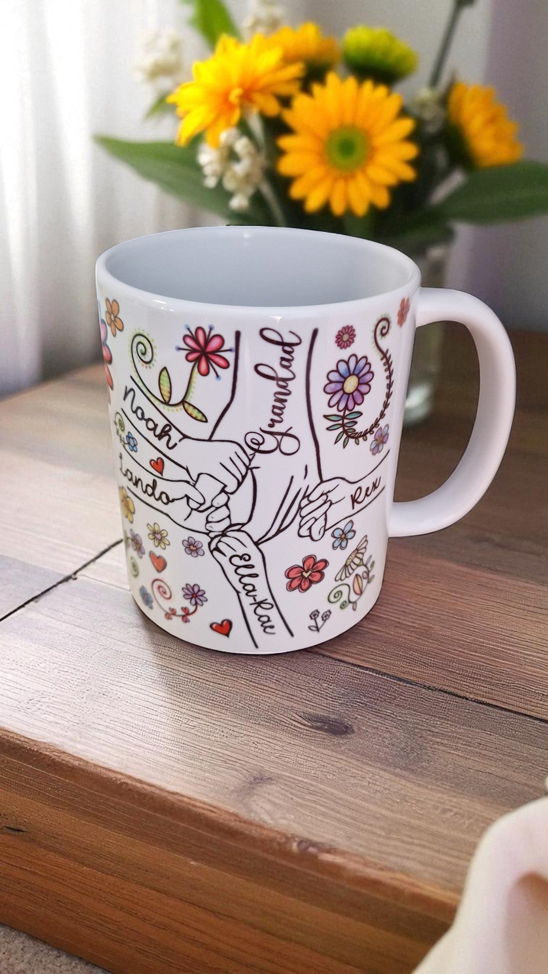 White mug with colorful designs and text on a wooden surface with flowers in the background