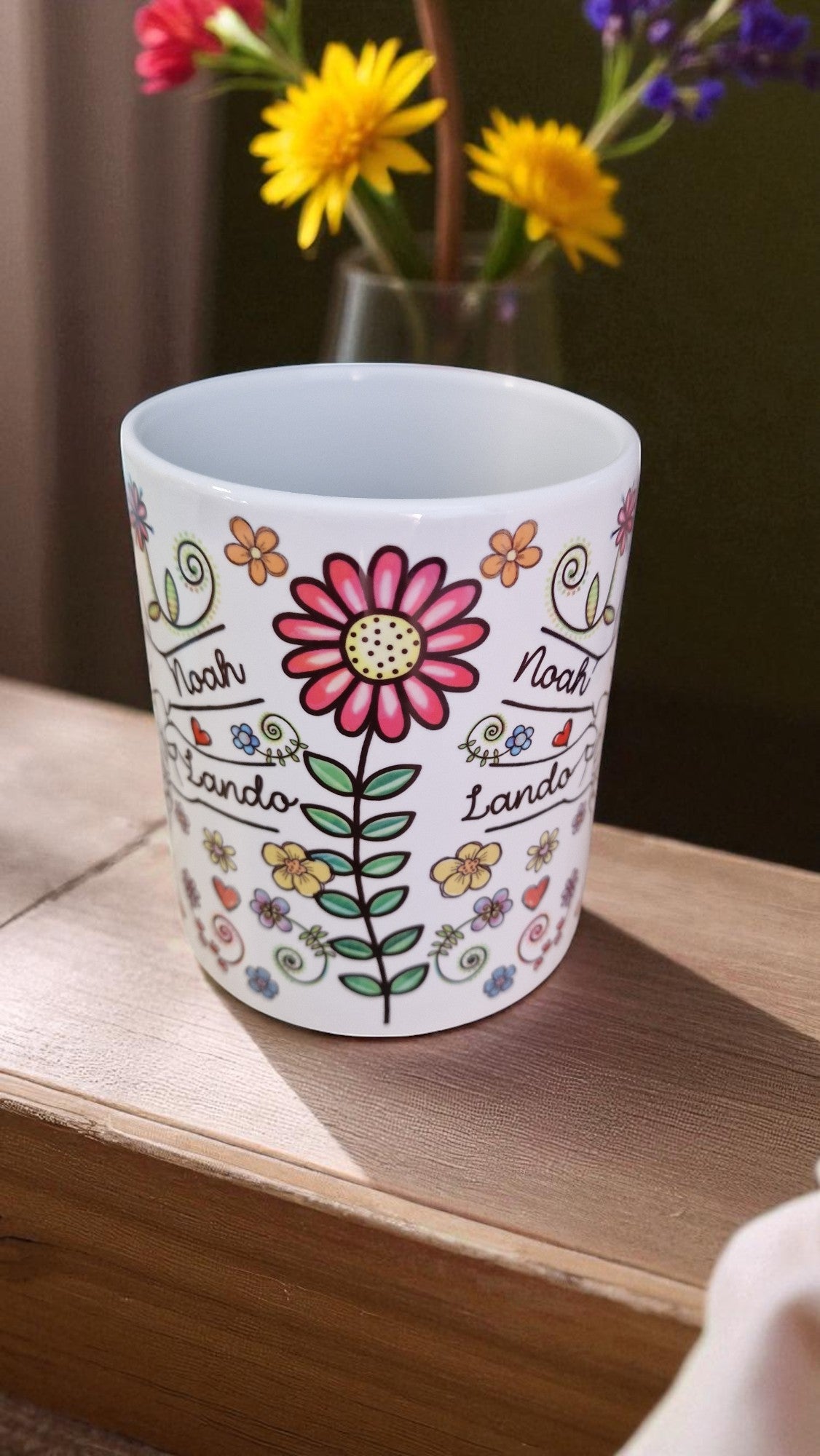Decorative mug with floral design and text on a wooden surface with flowers in the background