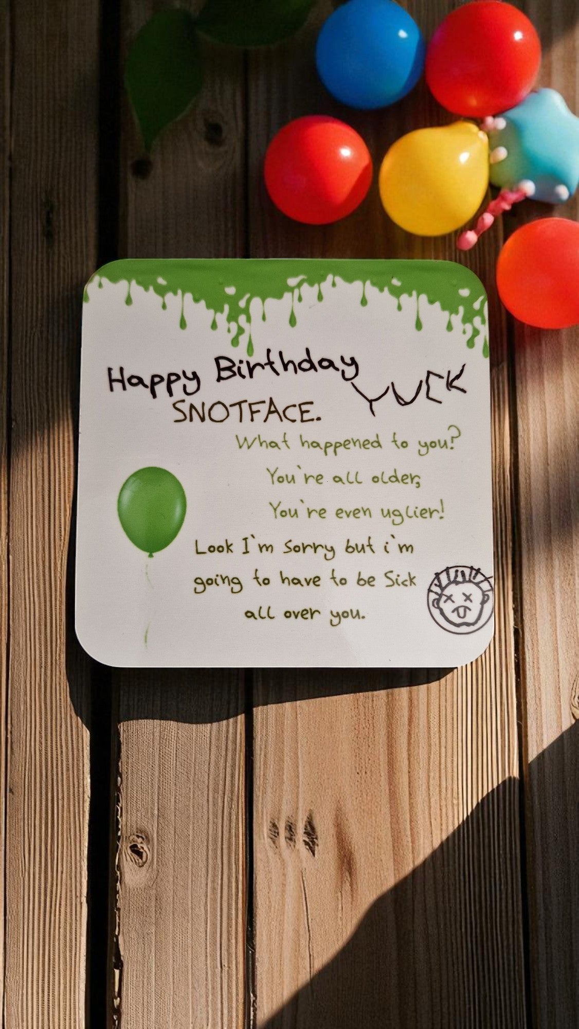 Square wooden coaster with green drips and the text “Happy Birthday Snotface. Yuck!” with a cartoon face and balloon, on a wooden table.