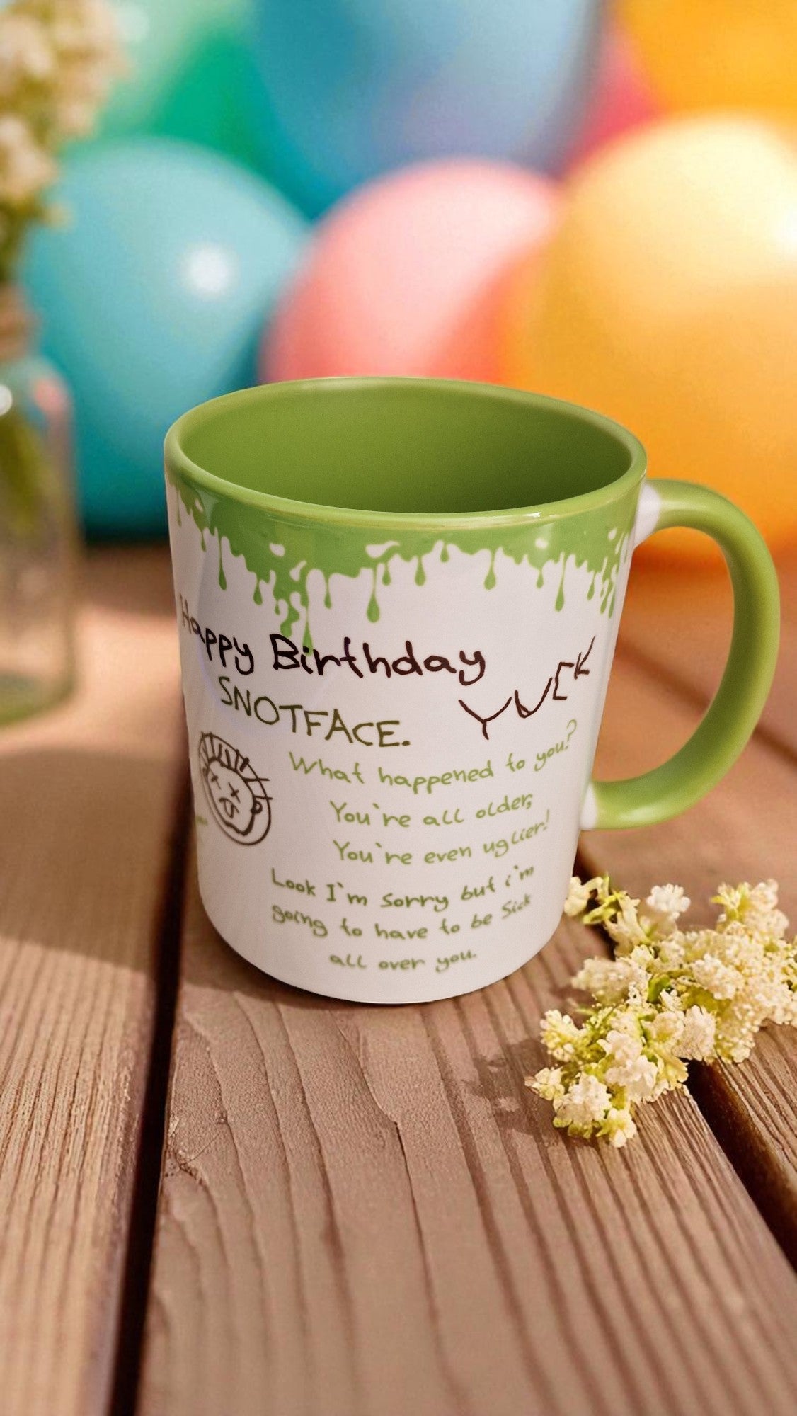 Green and white mug with humorous text on a wooden surface with balloons in the background