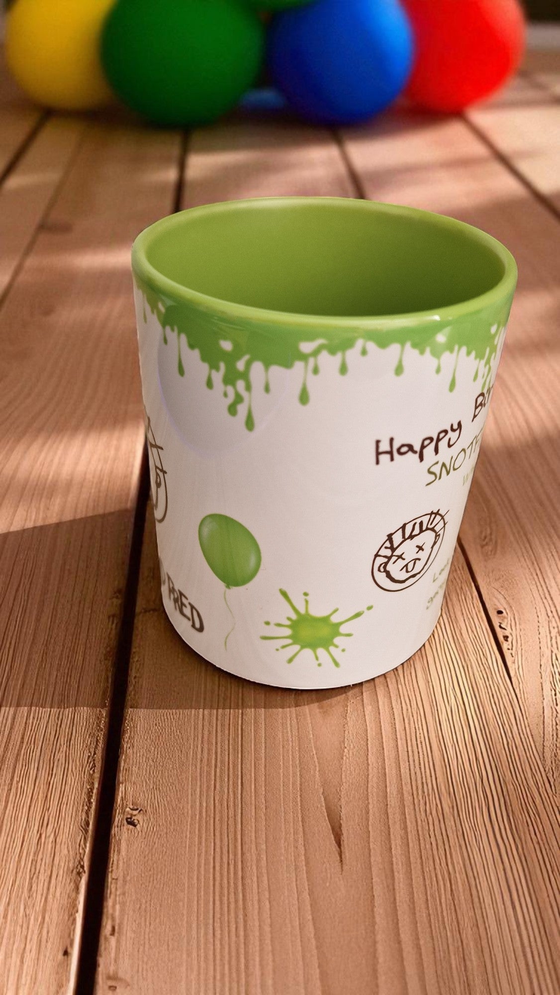 Green and white cup with text and graphics on a wooden surface with colorful balls in the background