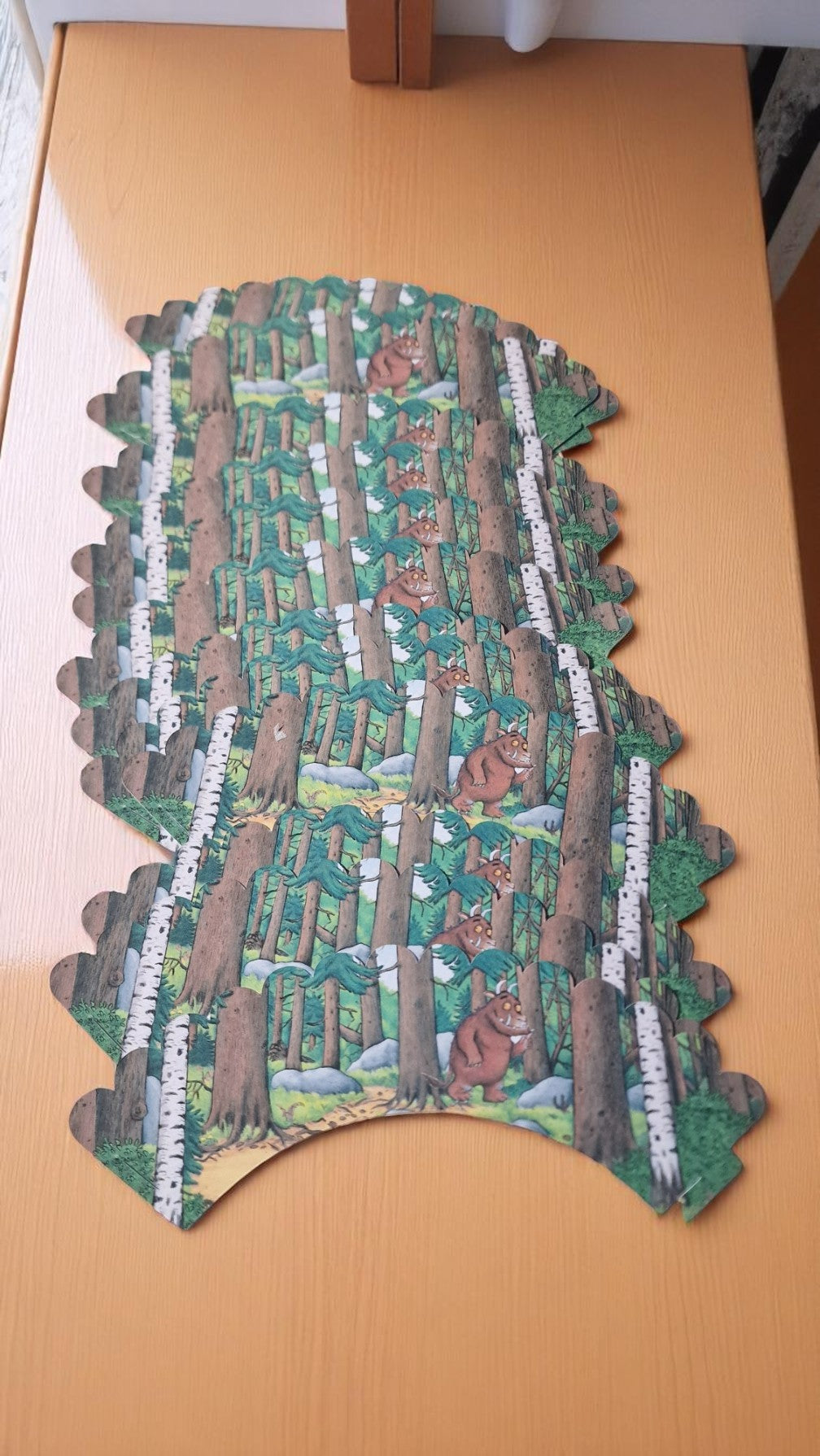 Gruffalo-themed cupcake case wrapper made of cardstock, featuring a colorful forest background with the Gruffalo character in the center, shown flat and unassembled on a wooden surface.