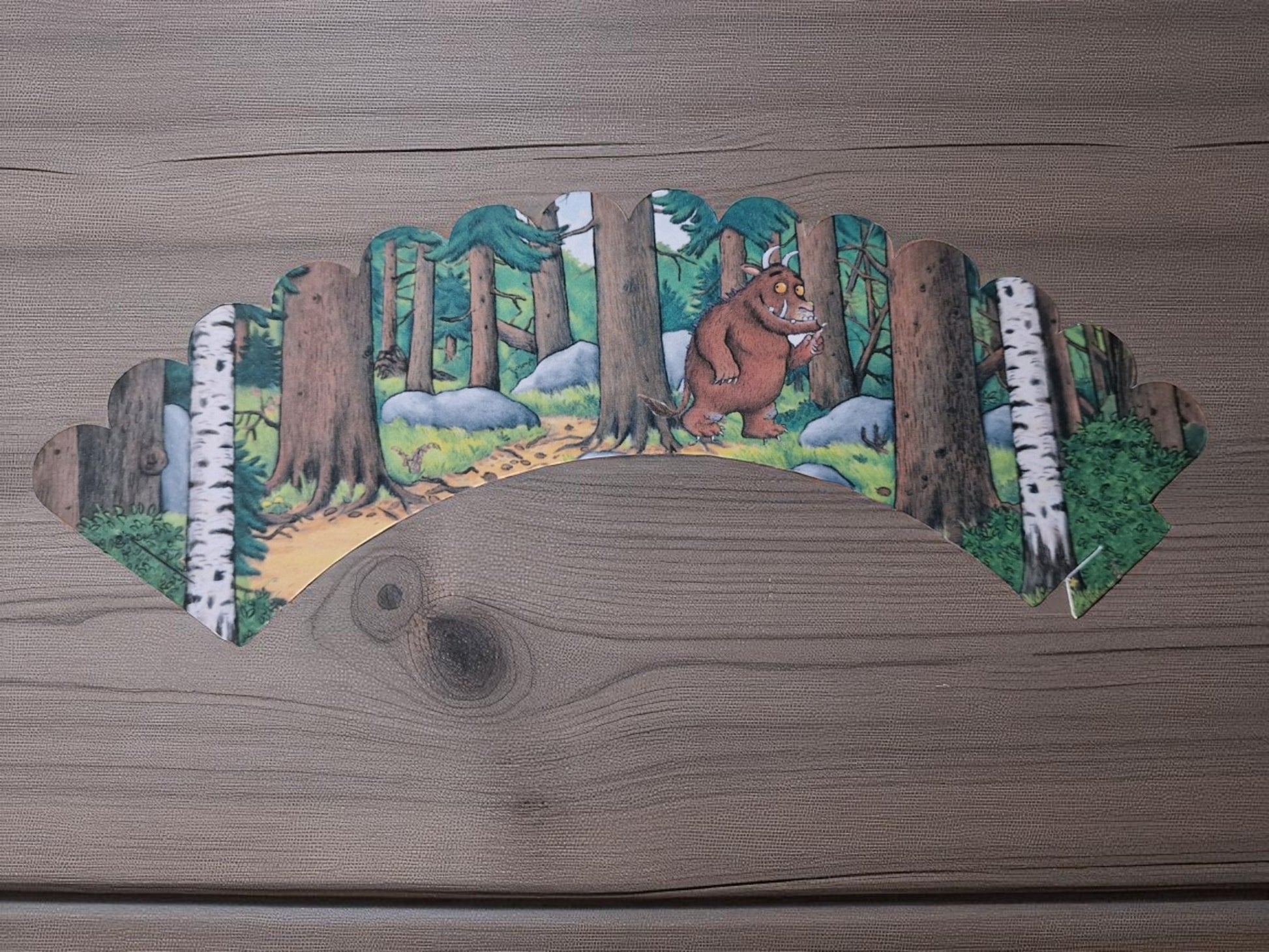 Gruffalo-themed cupcake case wrapper made of cardstock, featuring a colorful forest background with the Gruffalo character in the center, shown flat and unassembled on a wooden surface.