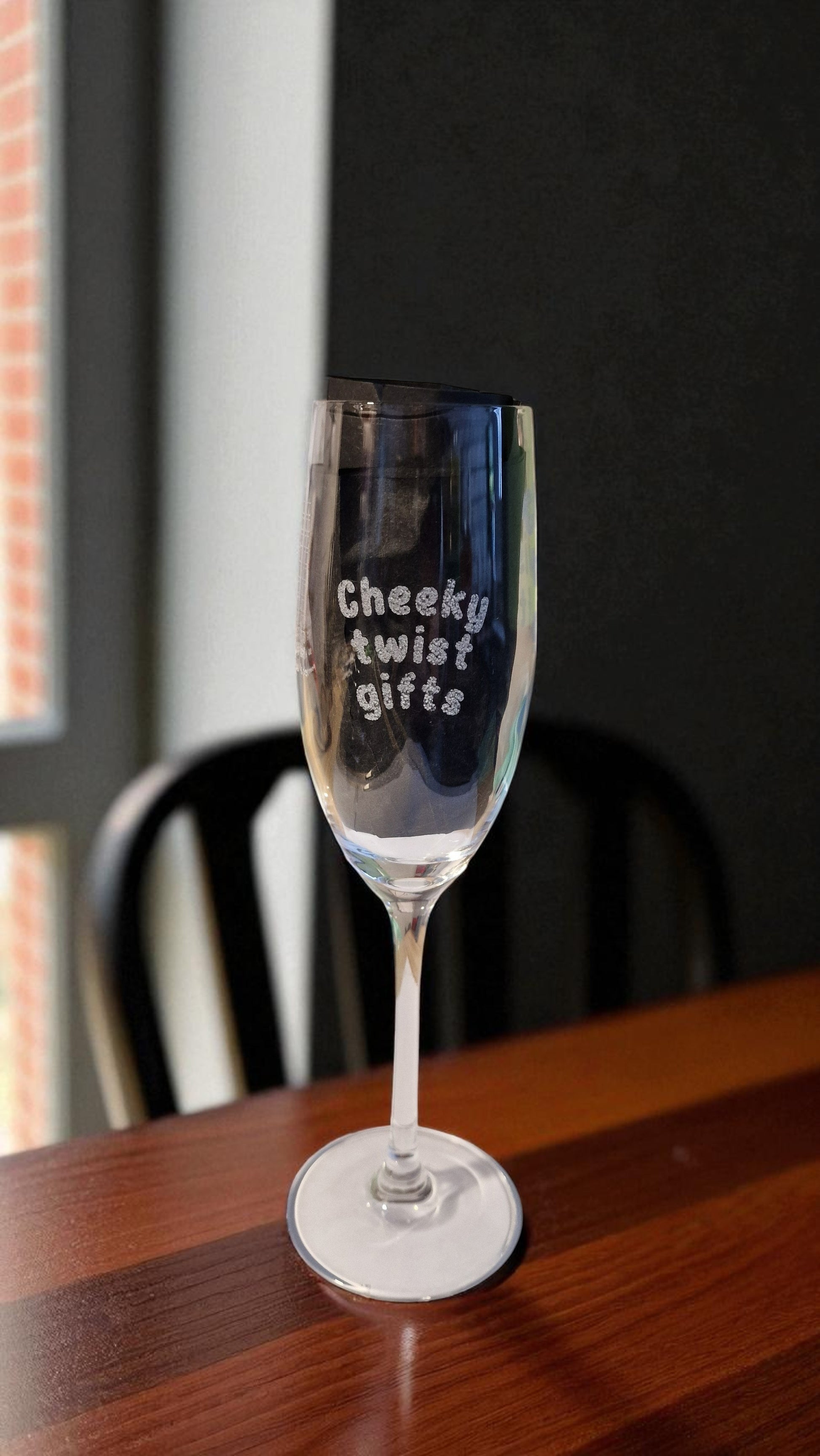 Personalised champagne flute glass engraved with ‘Cheeky twist gifts’, filled with rosé, on a marble kitchen counter with grapes and a bottle in the background