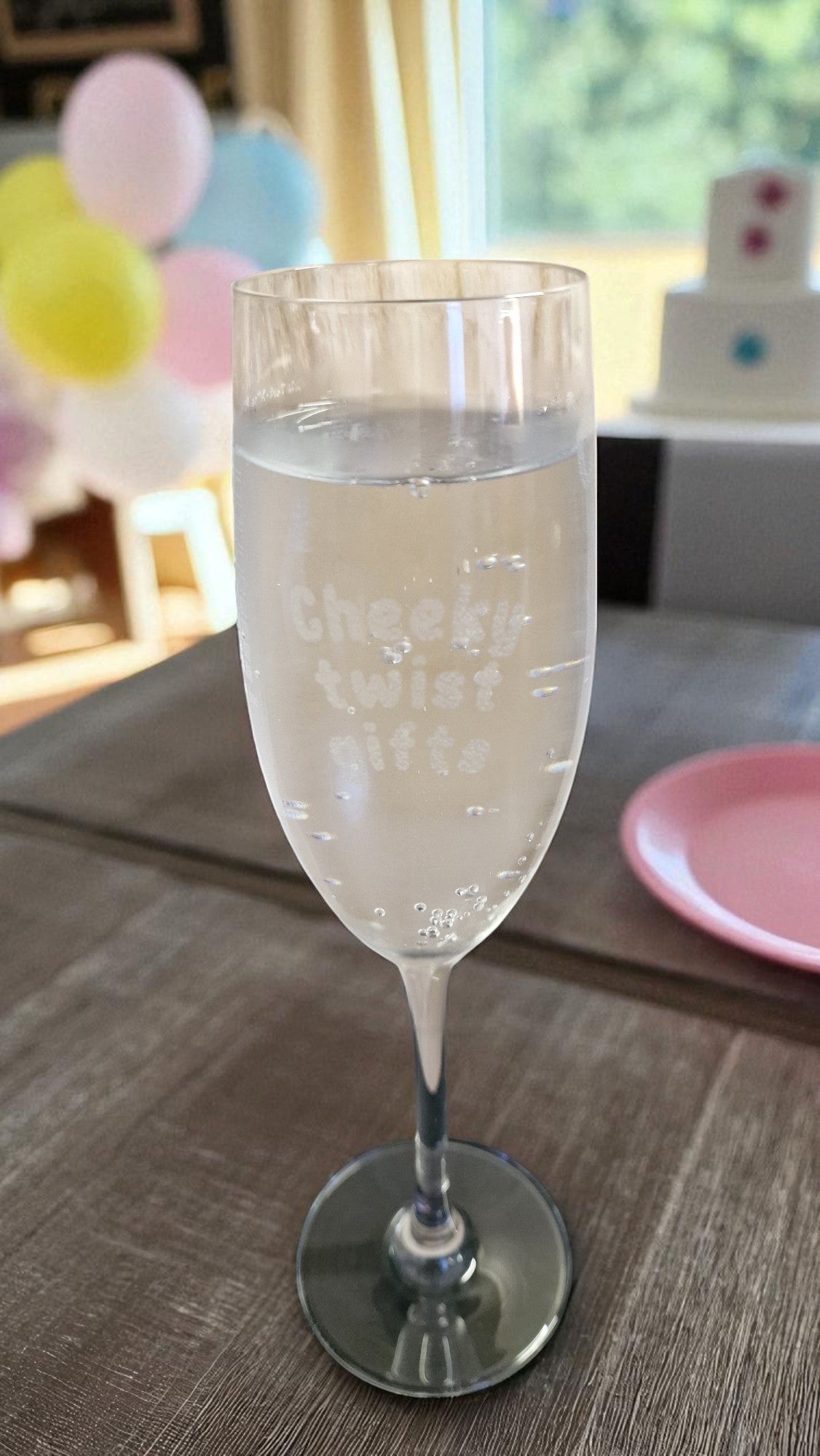 Personalised champagne flute glass engraved with ‘Cheeky twist gifts’, filled with rosé, on a marble kitchen counter with grapes and a bottle in the background