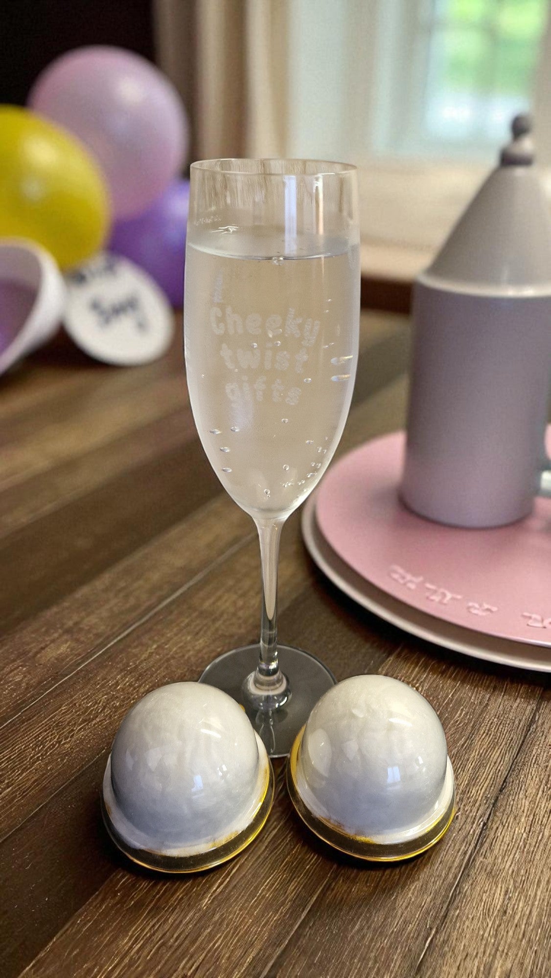 Personalised champagne flute glass engraved with ‘Cheeky twist gifts’, filled with rosé, on a marble kitchen counter with grapes and a bottle in the background