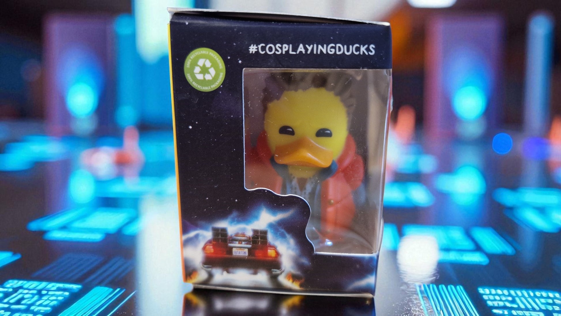 Packaged toy duck with '#CosplayingDucks' branding on a blurred futuristic background
