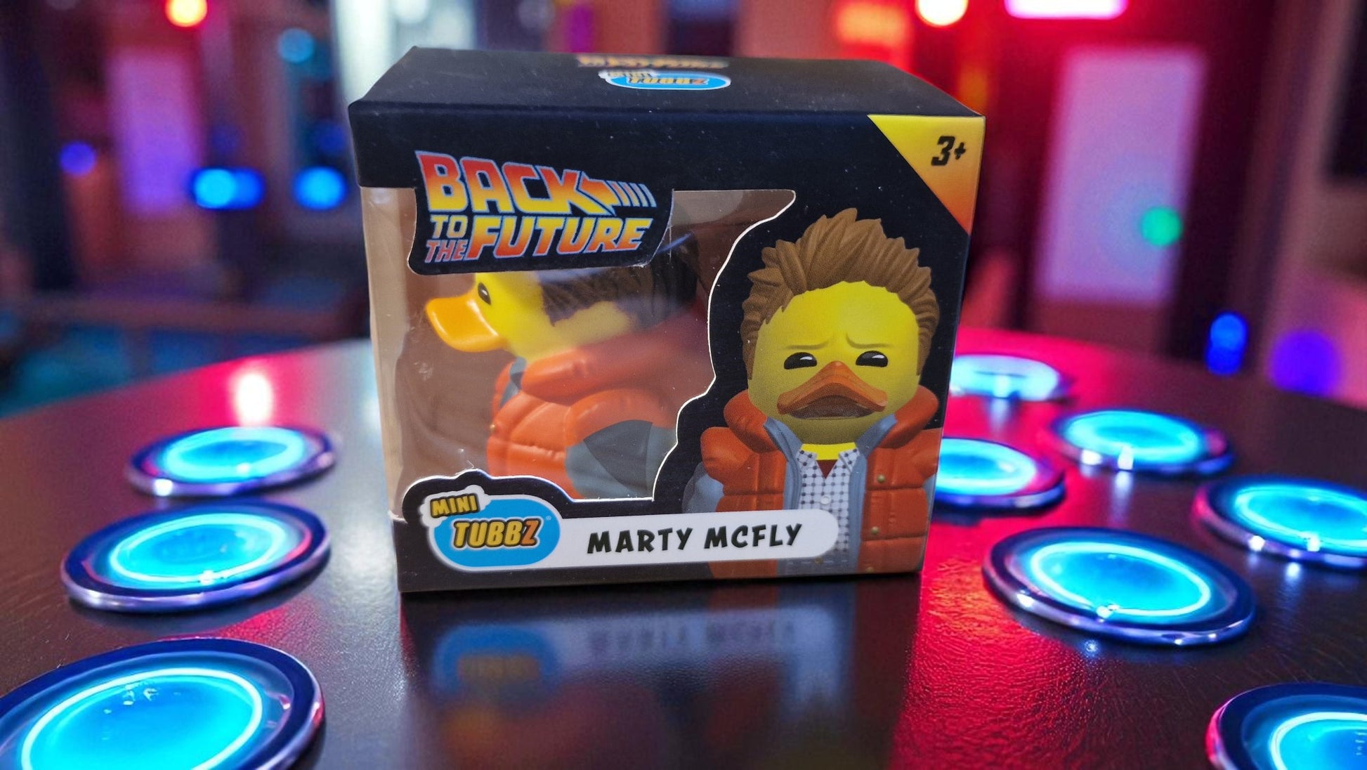 Toy figure of Marty McFly from 'Back to the Future' in packaging on a reflective surface with colorful lights in the background.