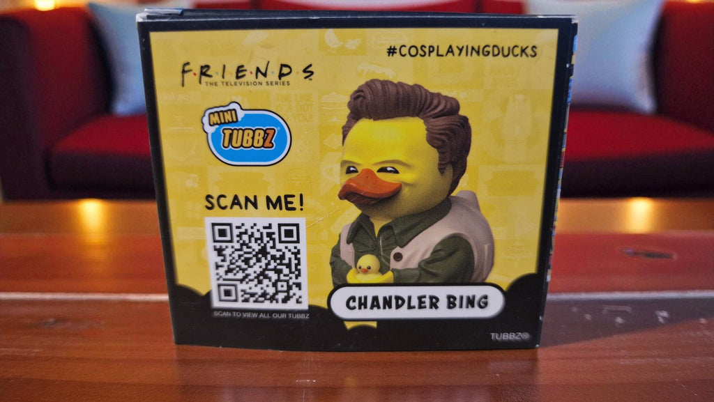 Rubber duck toy with 'Friends' character packaging featuring a QR code on a wooden surface.