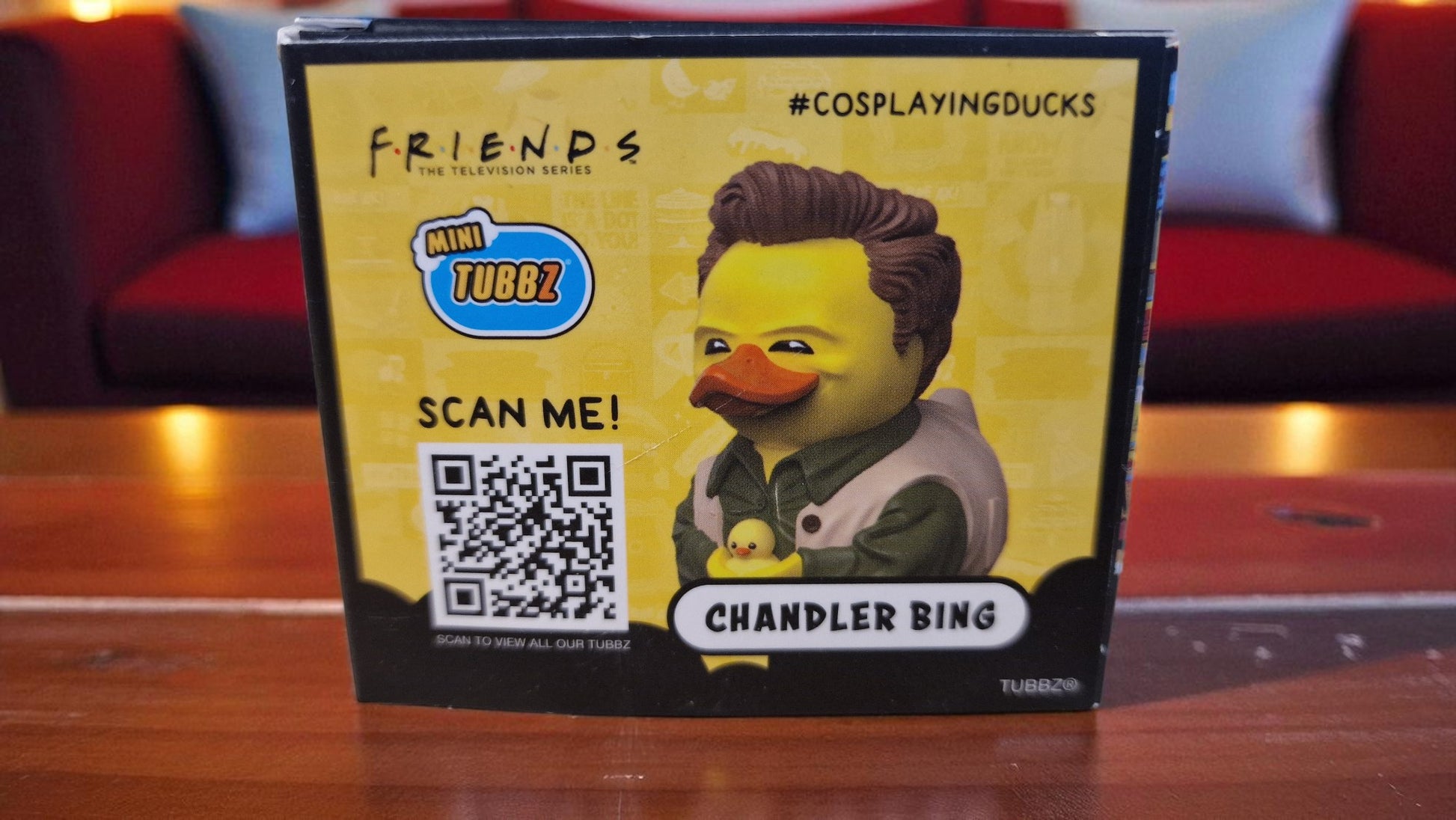 Rubber duck toy with 'Friends' character packaging featuring a QR code on a wooden surface.