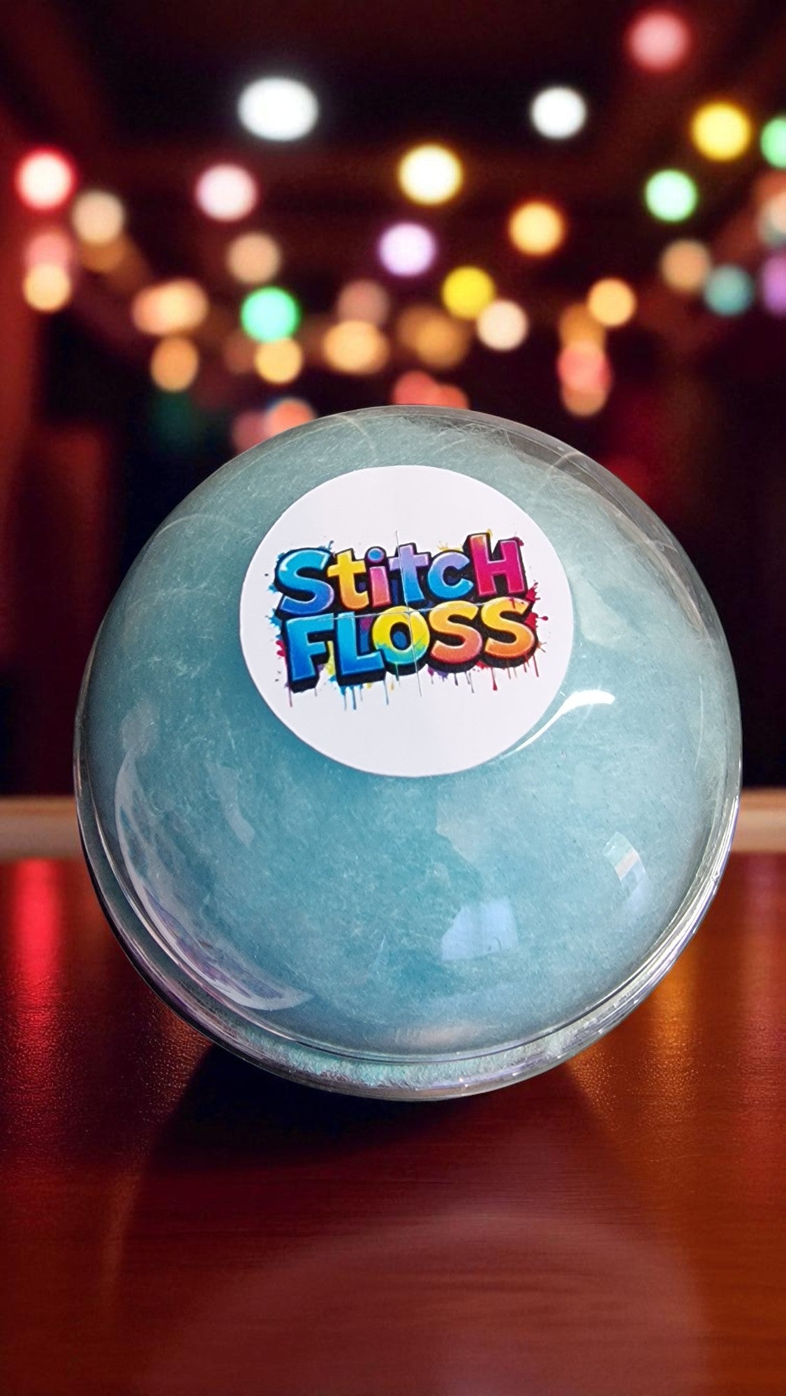 Ball of light blue yarn with a 'Stitch Floss' label on a wooden surface with colorful blurred lights in the background.