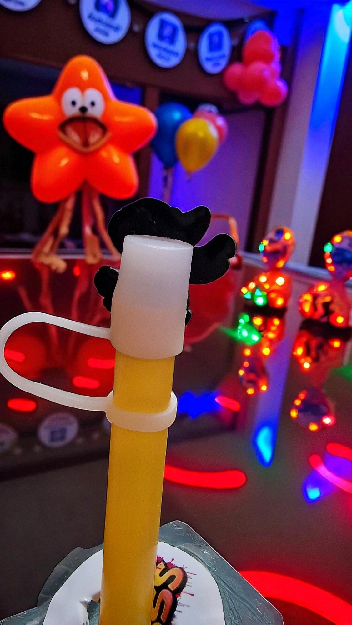 Toy with a yellow handle and black top in front of colorful balloons and lights.