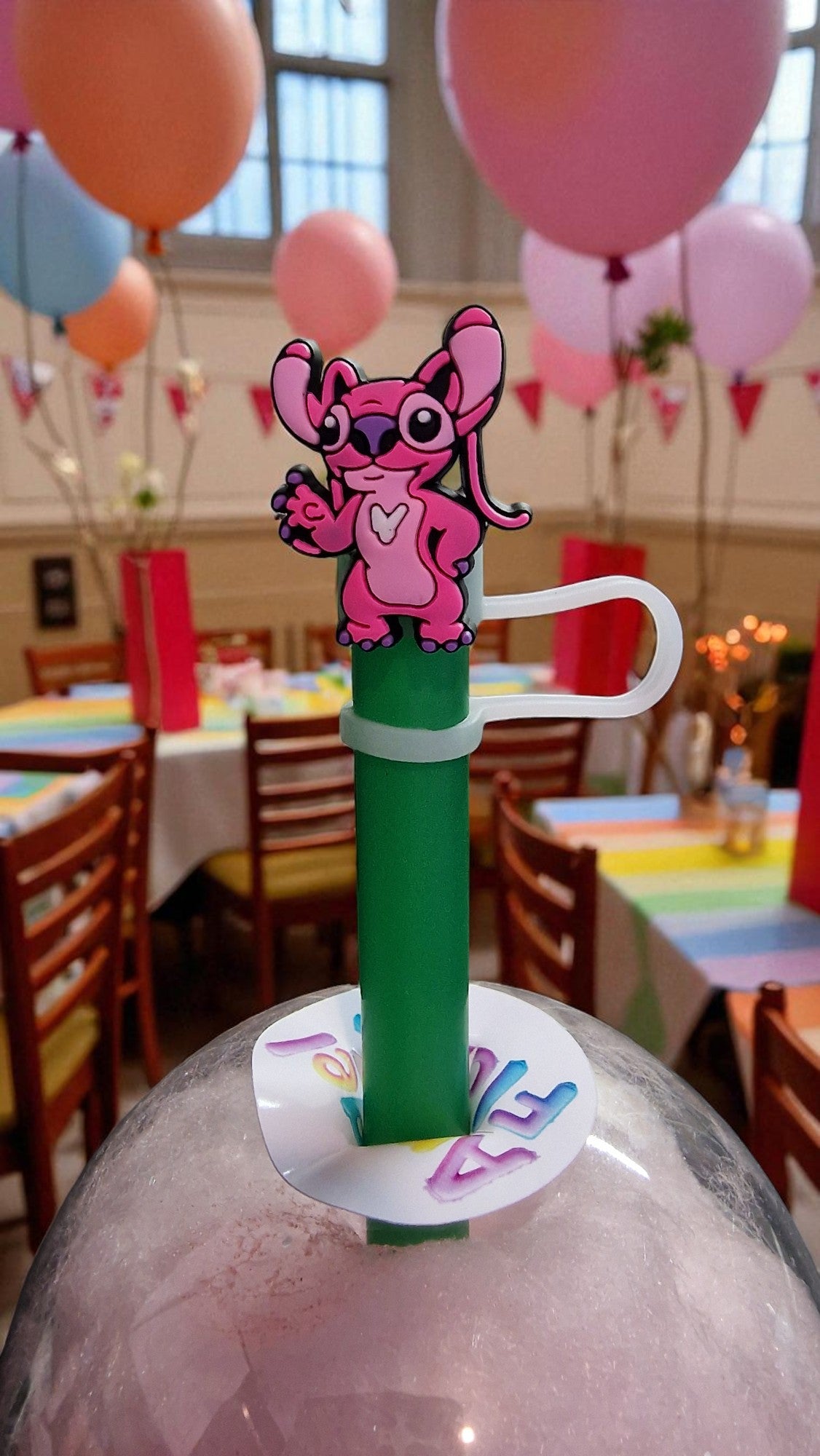 Pink cartoon character on a green stand with balloons and tables in the background