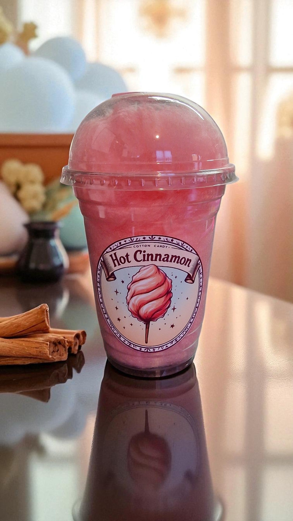 Pink drink in a clear cup with a lid, labeled 'Hot Cinnamon', on a reflective surface.