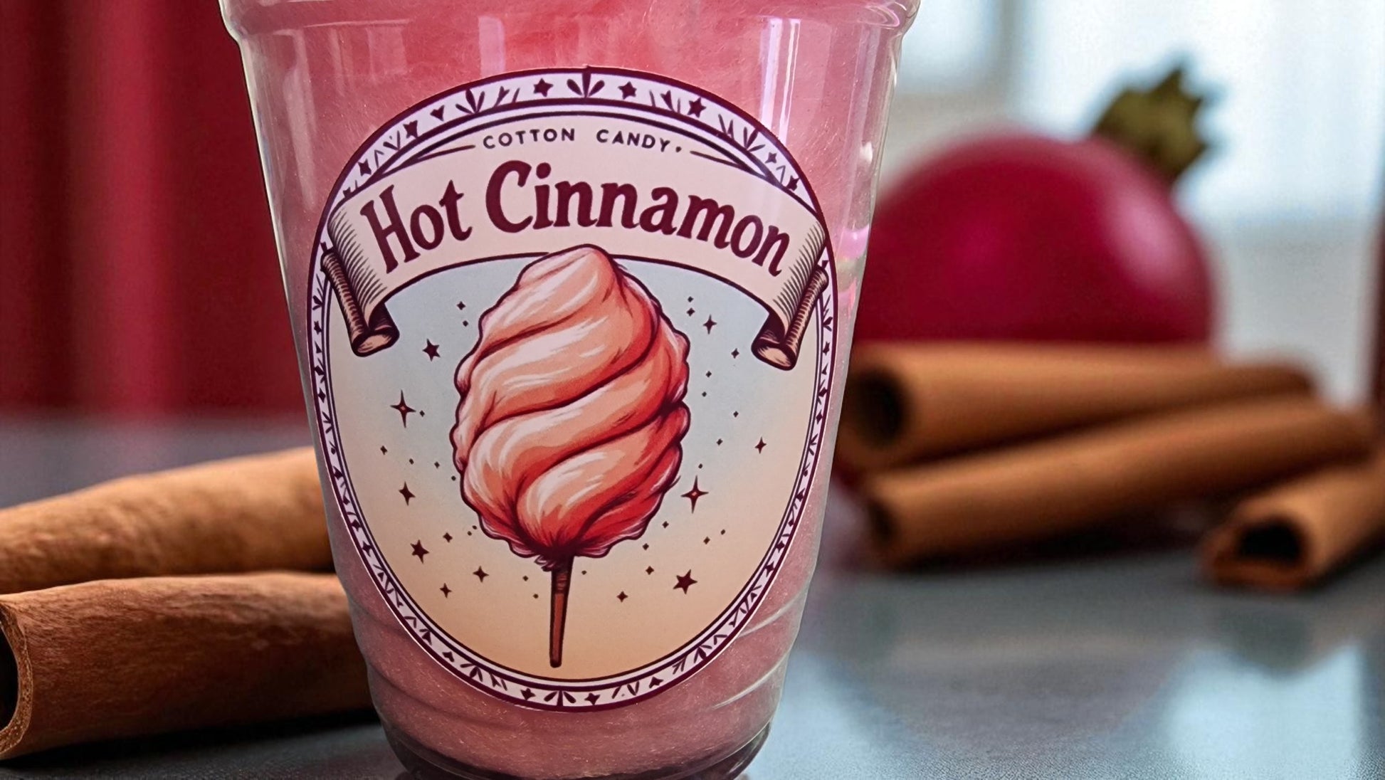 Pink drink cup labeled 'Hot Cinnamon' with cotton candy design, surrounded by cinnamon sticks and an apple.