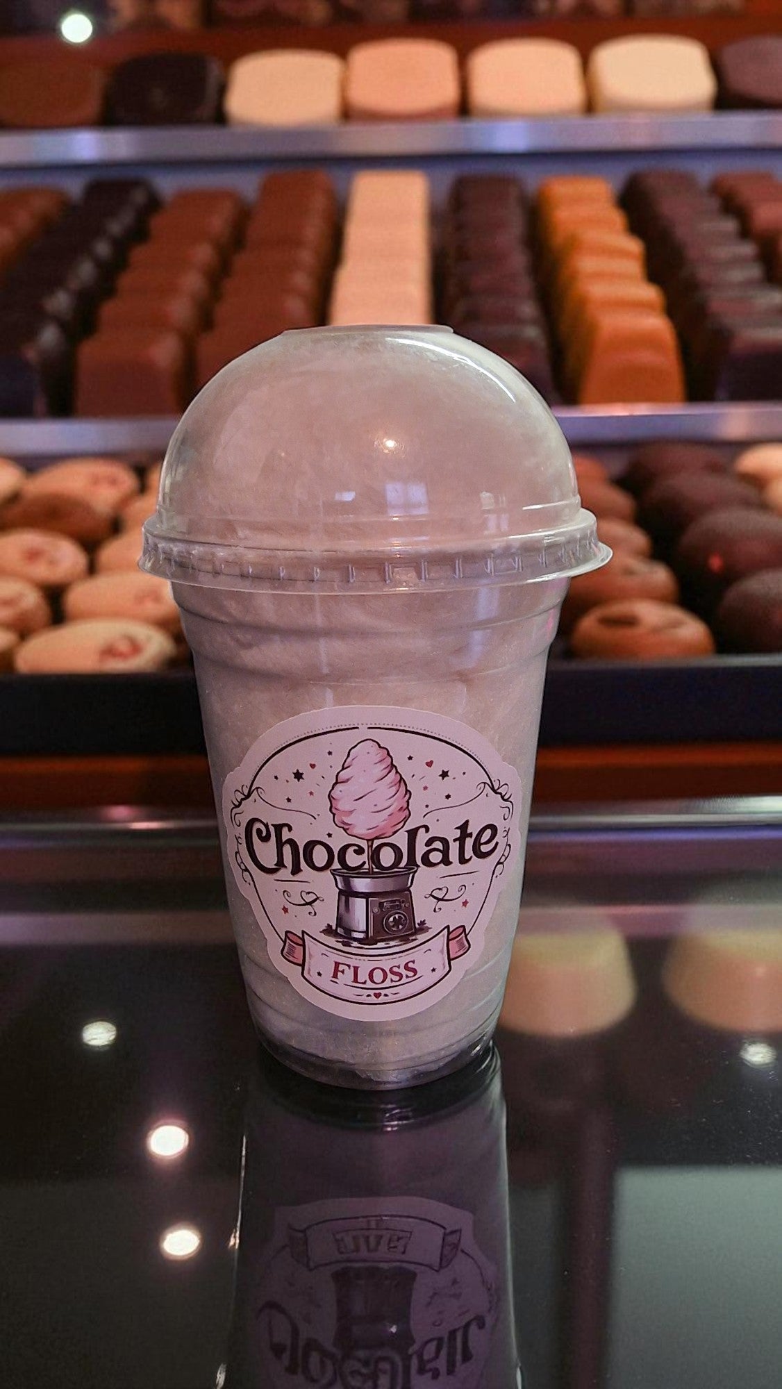 Ice cream cone-shaped cup with 'Chocolate Floss' branding in front of a display of chocolate-covered treats.
