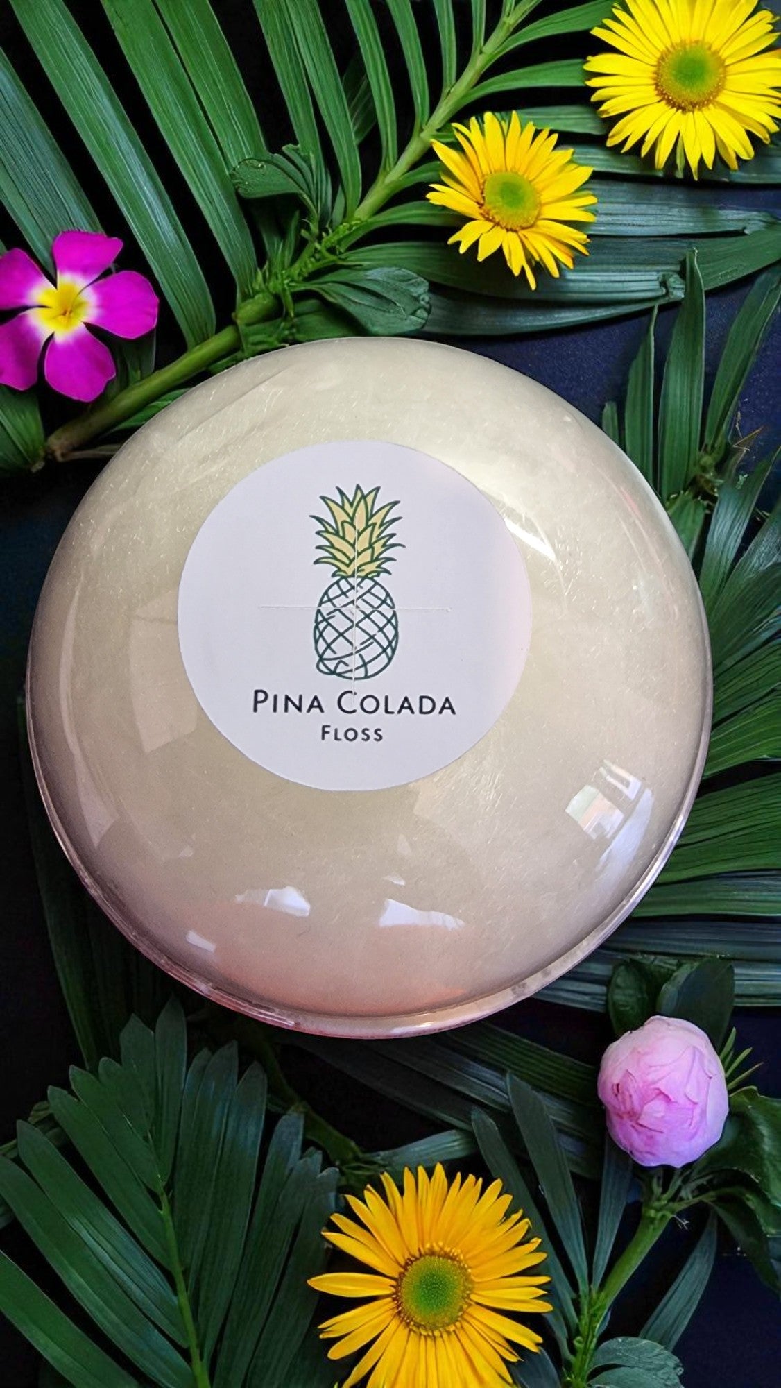 Round beige container labeled 'Pina Colada Floss' surrounded by tropical flowers and leaves.