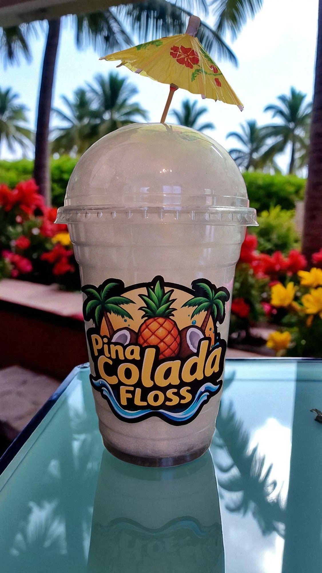 Pina Colada Floss drink with a pineapple label and umbrella on a reflective surface with blurred outdoor background.