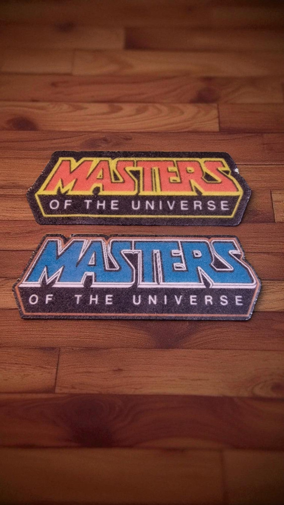 Two 'Masters of the Universe' patches on a wooden surface