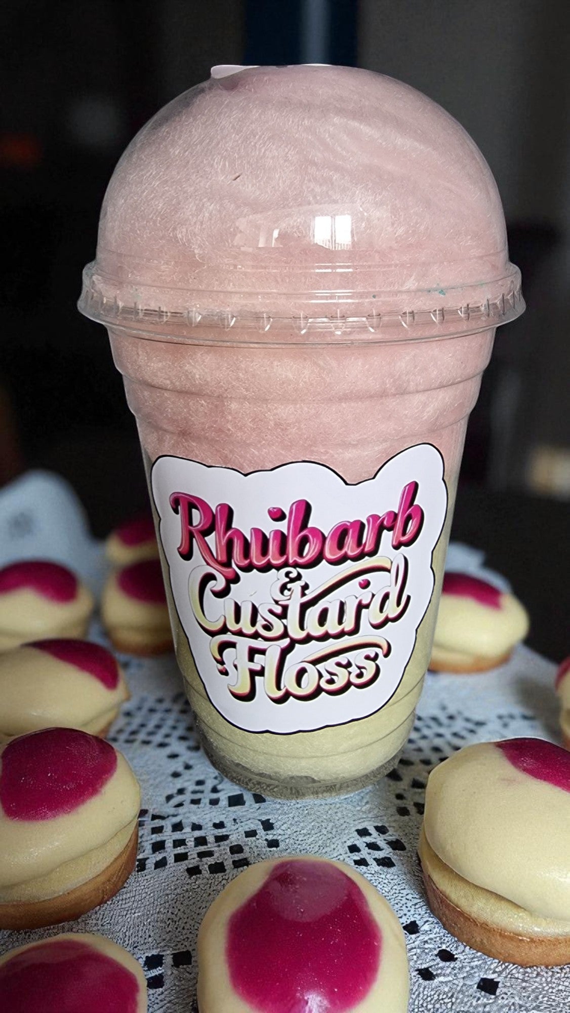 Pink shake in a plastic cup with 'Rhubarb Custard Floss' label, surrounded by cookies on a lace tablecloth.
