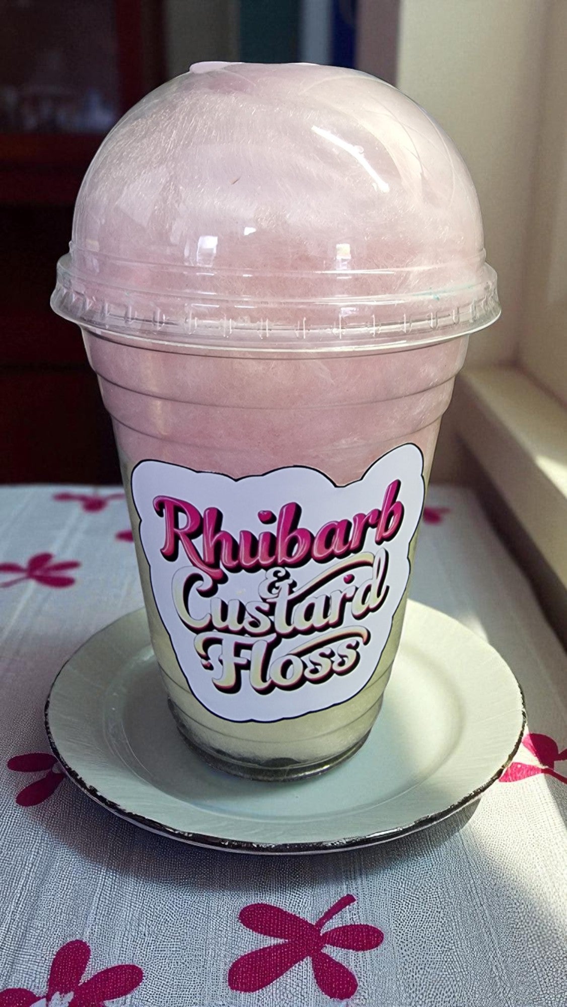 Pink shake in a plastic cup with a 'Rhubarb Custard Floss' label on a floral tablecloth.